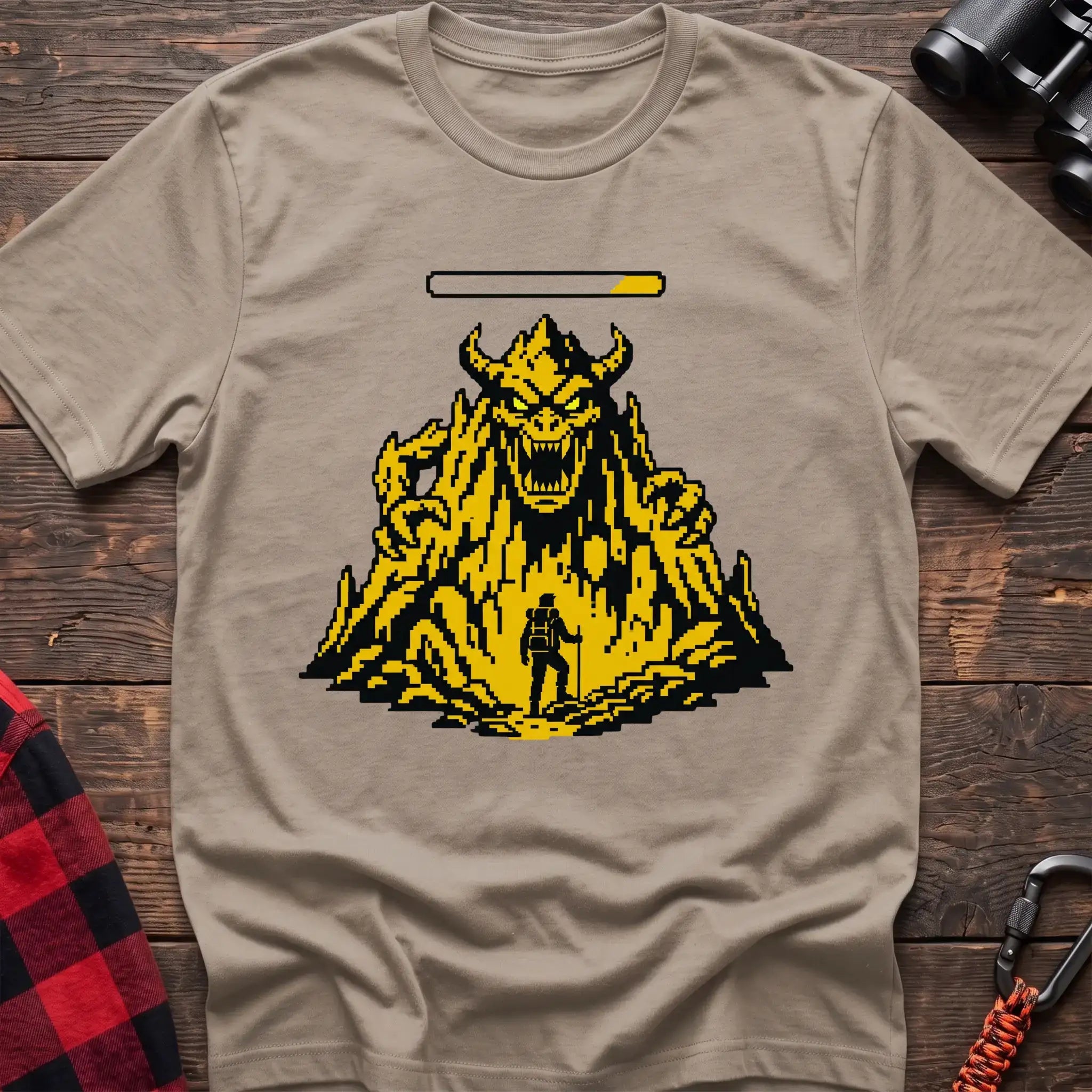 Sand t-shirt with a pixel-art illustration of a hiker facing a large horned mountain monster with a health bar above it, laid flat on a wooden surface.