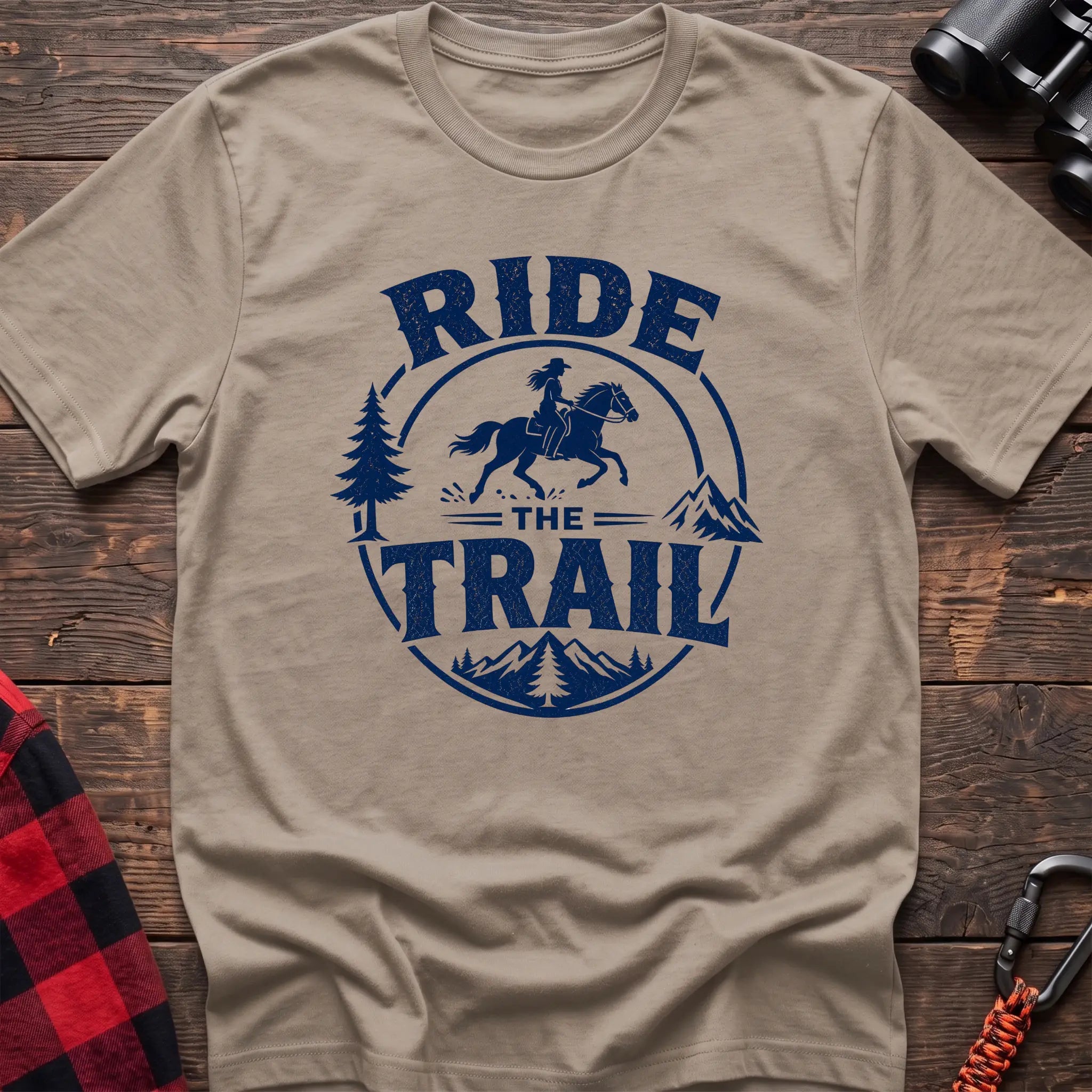 Sand cotton t-shirt with navy Ride the Trail graphic of a woman horseback riding through mountains and pine trees, flat lay on rustic wood with hiking gear