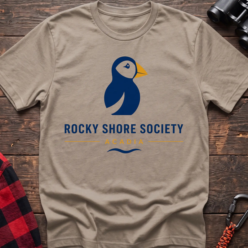 Sand cotton t-shirt with a navy puffin logo, yellow beak, and Rocky Shore Society Acadia text on a rustic wood background