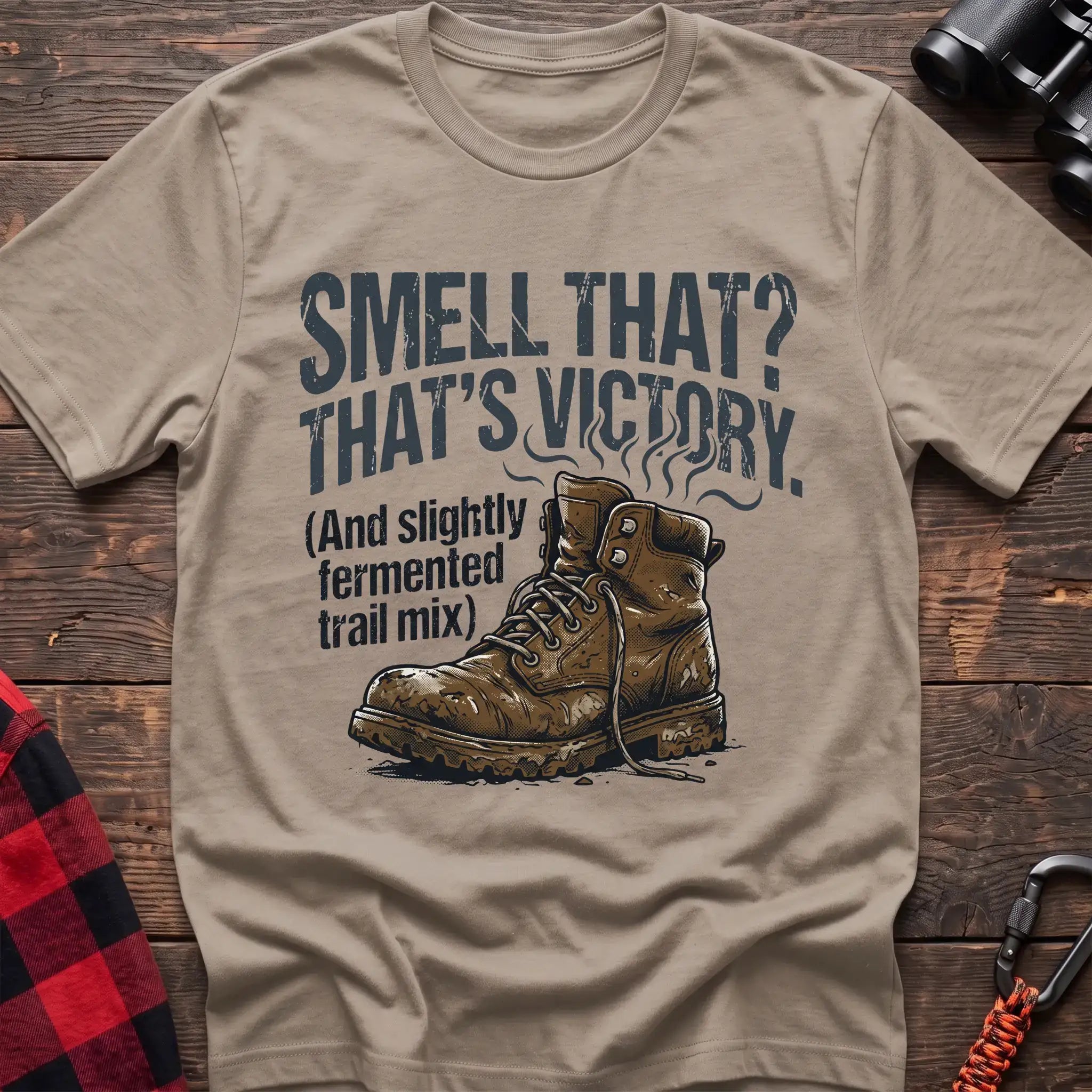 Sand t-shirt with an illustration of a dirty hiking boot with rising steam lines, and the text “SMELL THAT? THAT’S VICTORY. (And slightly fermented trail mix),” laid flat on a wooden surface