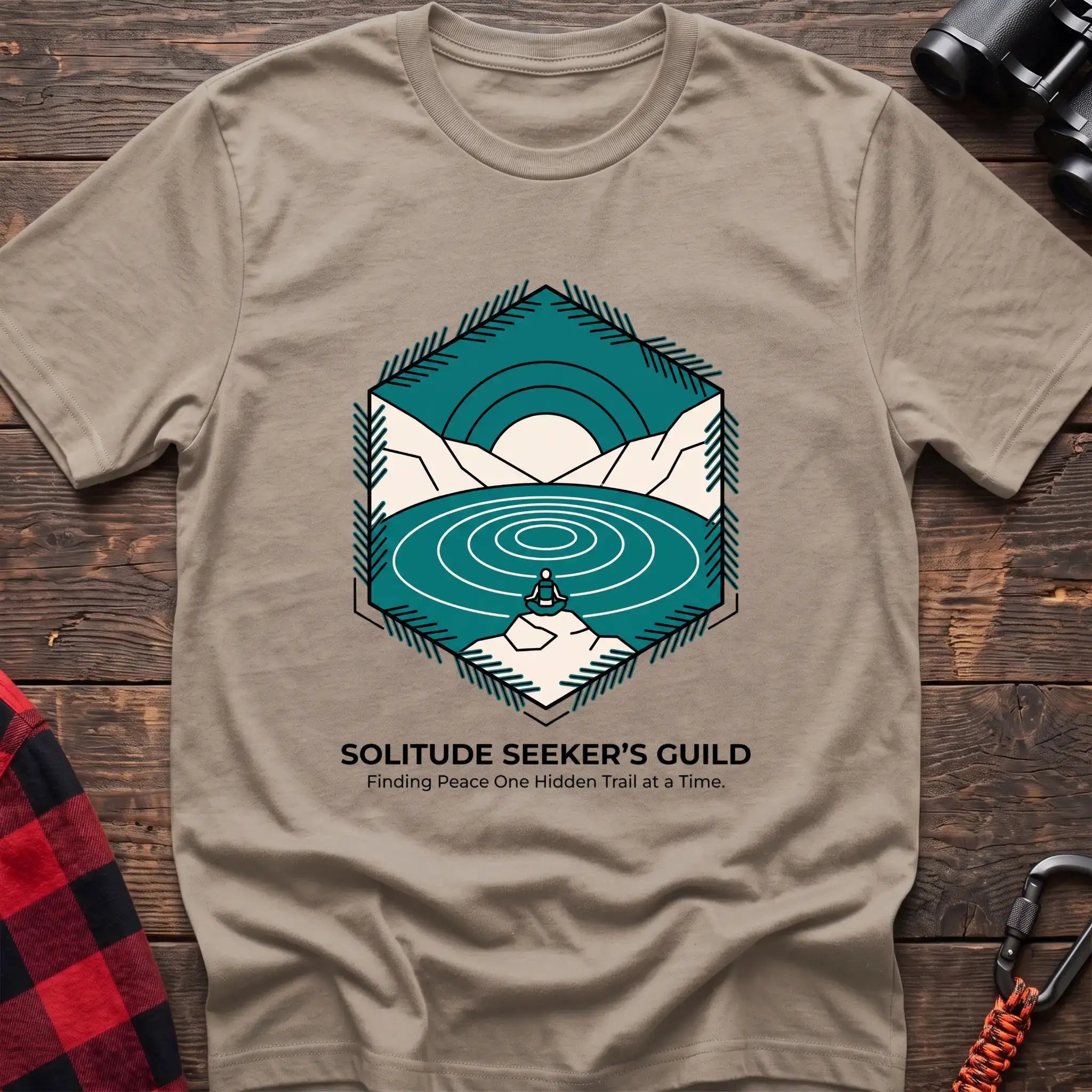 Sand t-shirt with a hexagonal illustration of a person meditating by a mountain lake, with the text “SOLITUDE SEEKER’S GUILD” and “Finding Peace One Hidden Trail at a Time,” laid flat on a wooden surface.