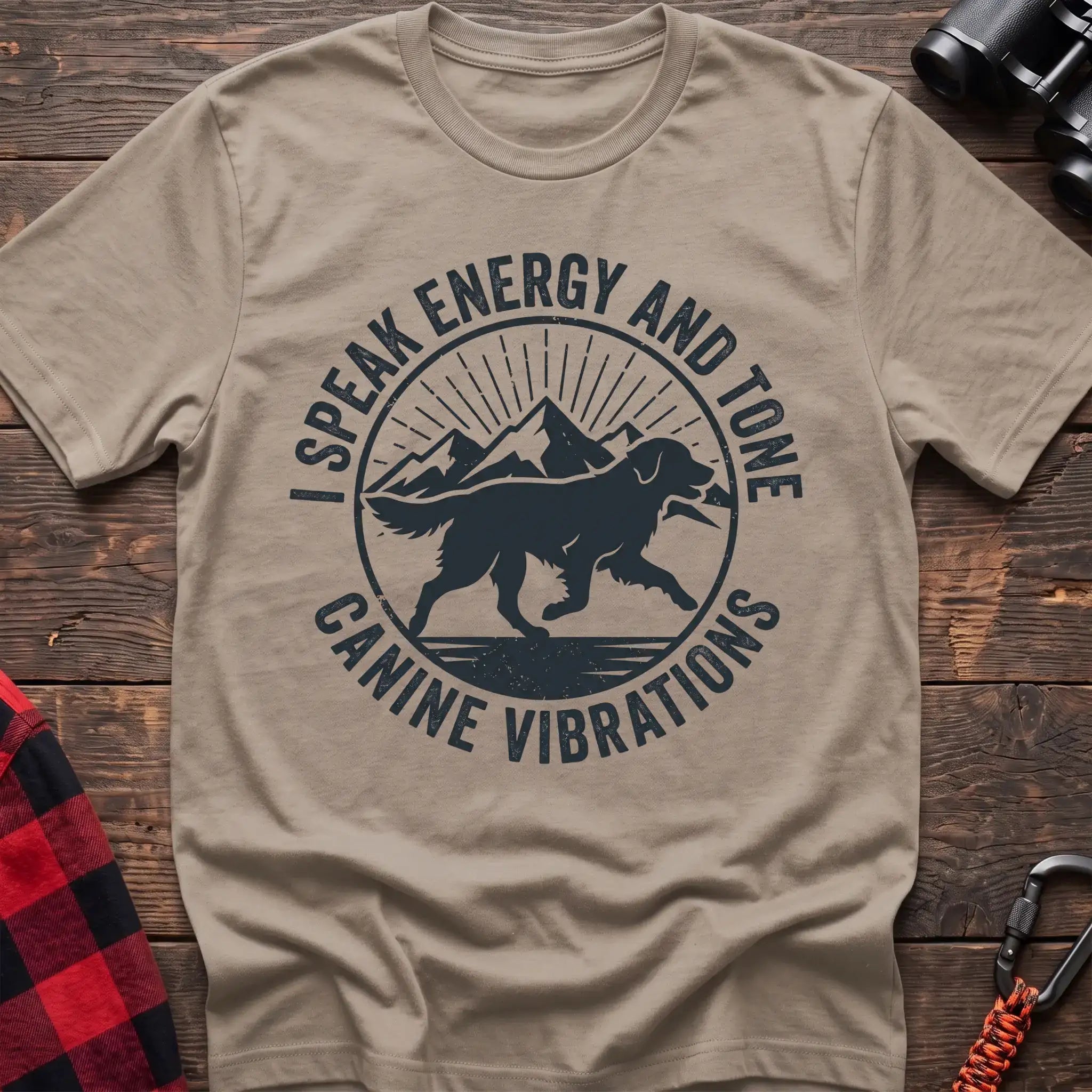 Sand t-shirt with a circular dark gray badge design featuring a running dog carrying a stick against mountain peaks and sun rays, with the text “I SPEAK ENERGY AND TONE” and “CANINE VIBRATIONS,” laid flat on a wooden surface.