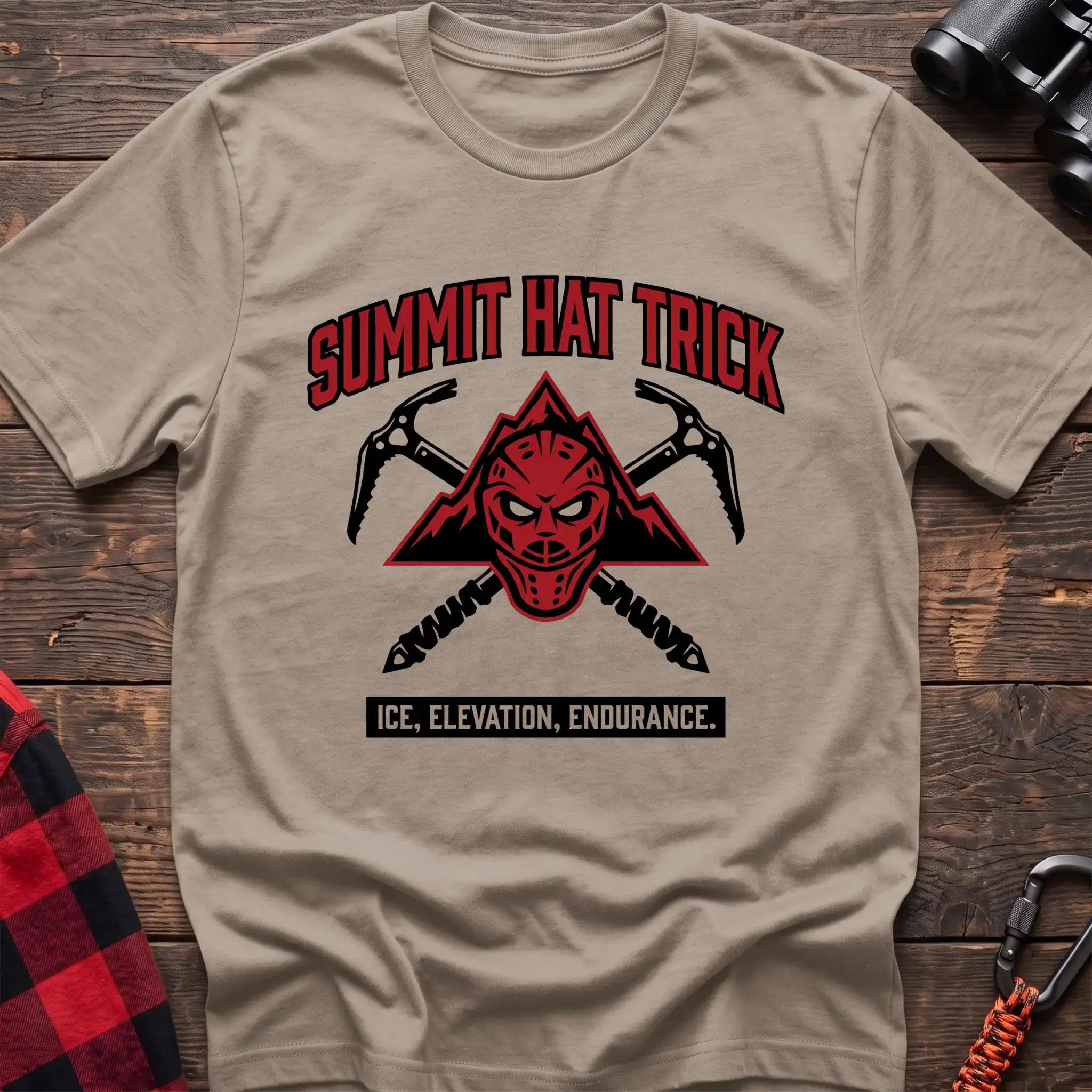 Sand t-shirt with a red mountain-themed goalie mask and crossed ice axes, with the text “SUMMIT HAT TRICK” and “ICE, ELEVATION, ENDURANCE,” laid flat on a wooden surface.