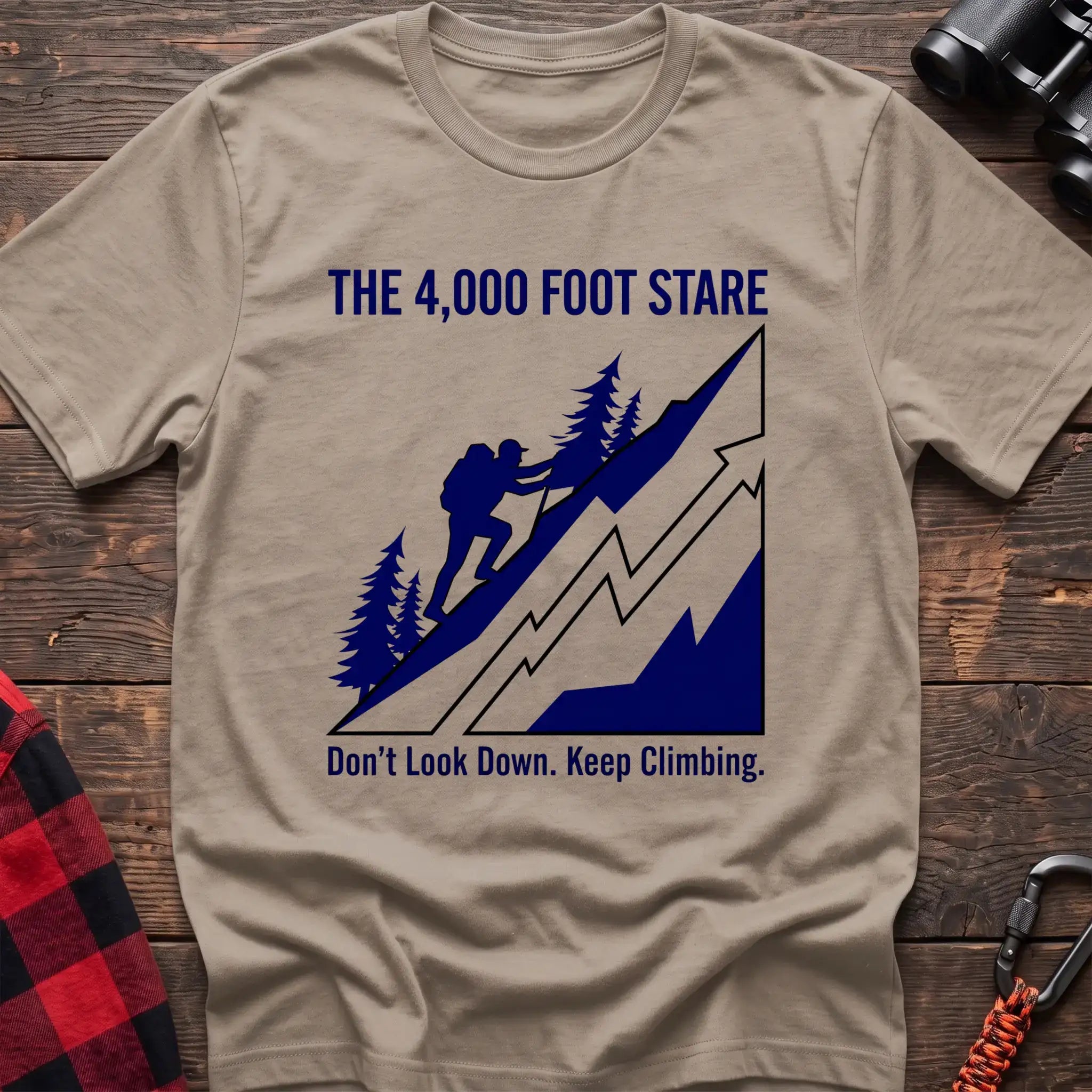 Sand t-shirt with a blue illustration of a hiker climbing a steep mountain, with the text “THE 4,000 FOOT STARE” and “Don’t Look Down. Keep Climbing,” laid flat on a wooden surface.