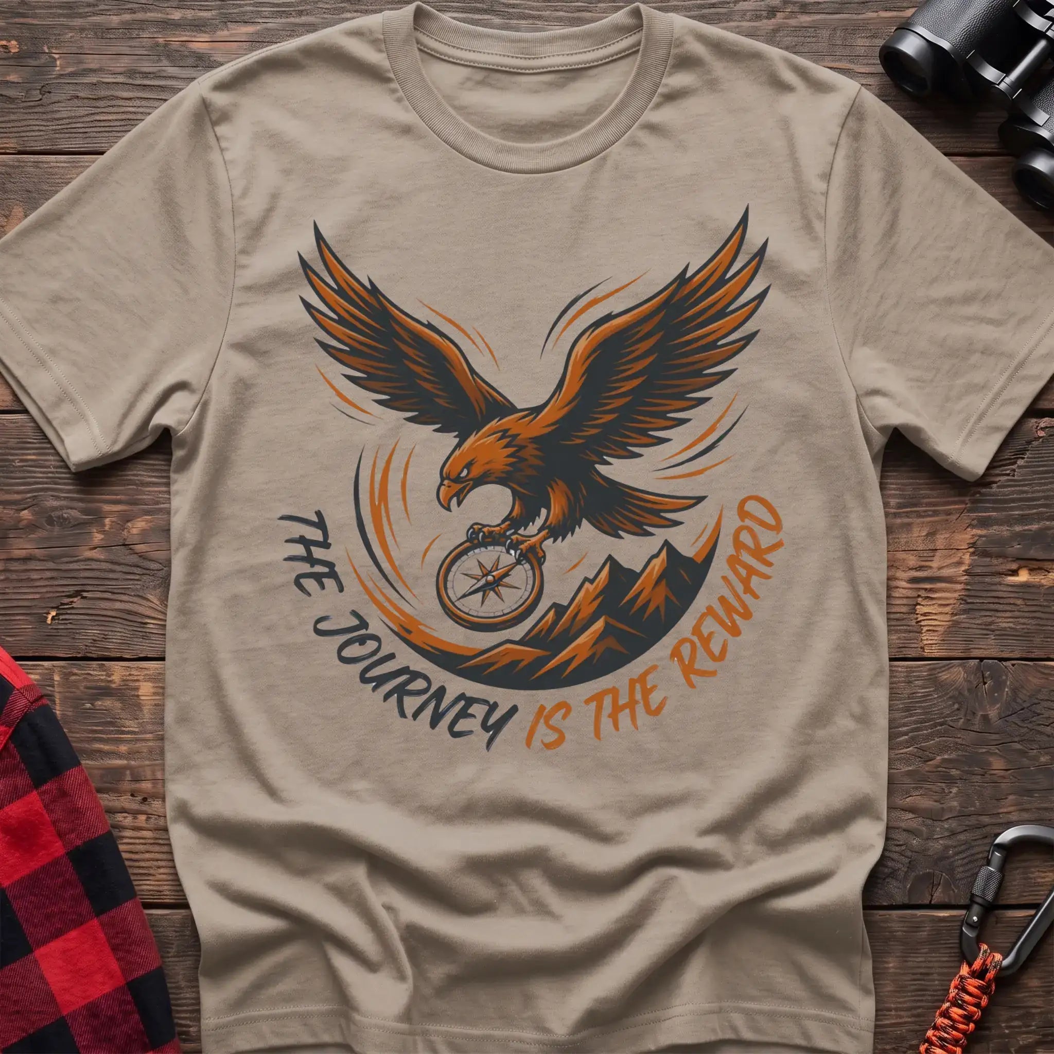 Sand t-shirt with an orange and dark graphic of a soaring eagle holding a compass above mountains, with the curved text “THE JOURNEY IS THE REWARD,” laid flat on a wooden surface.