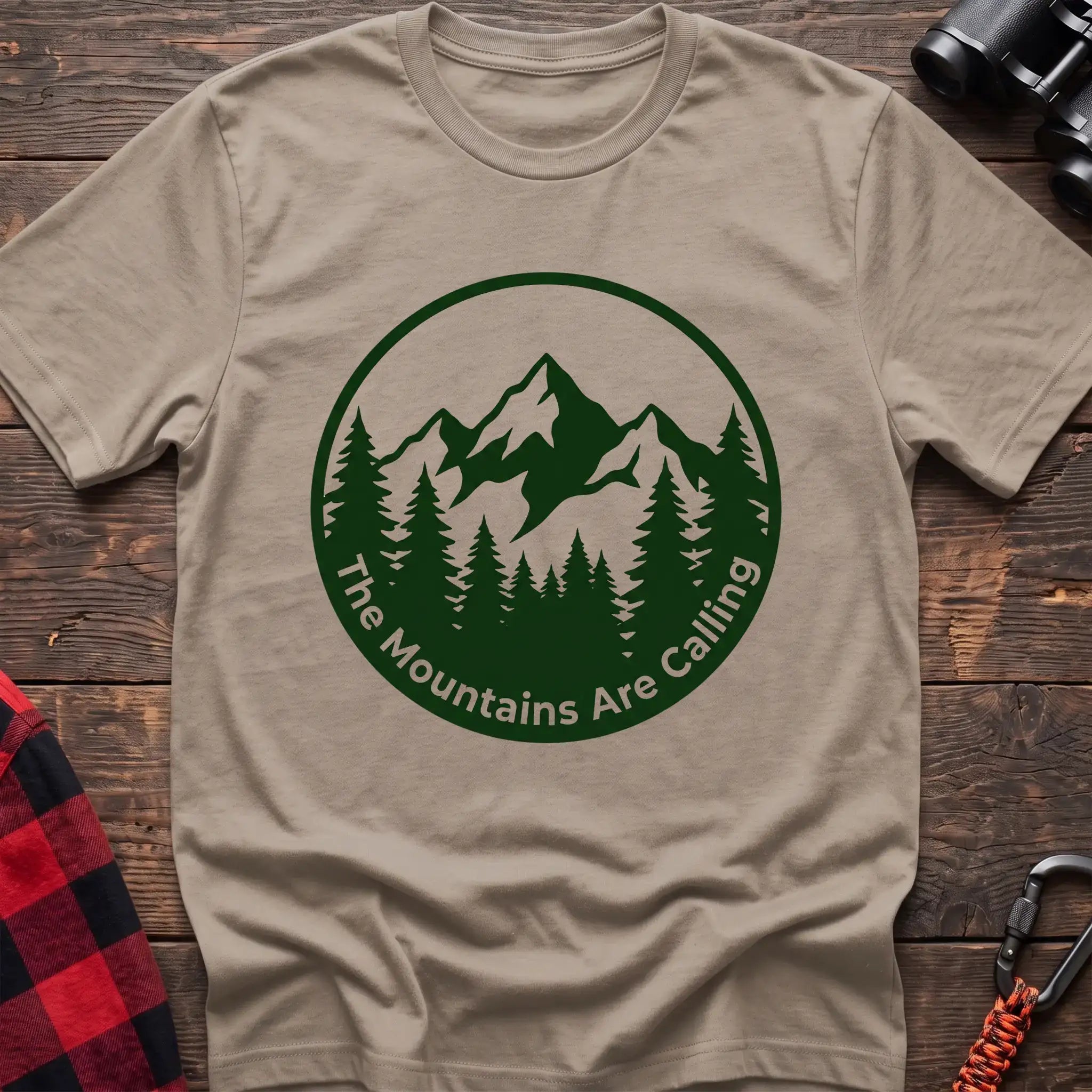 Sand t-shirt with a circular dark green design featuring mountain peaks and pine trees, with the text “The Mountains Are Calling,” laid flat on a wooden surface.