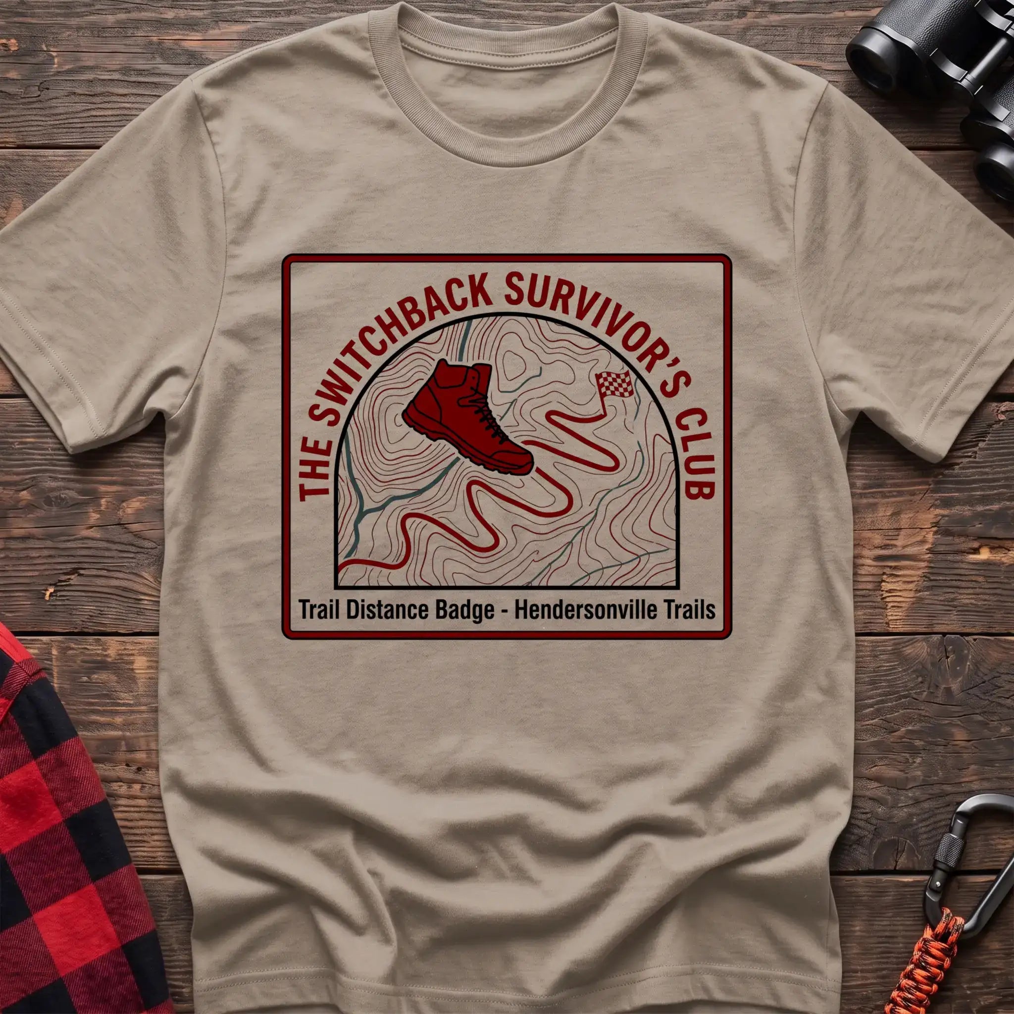 Sand t-shirt with a square badge design featuring a red hiking boot on a topographic map with a winding trail, and the text “THE SWITCHBACK SURVIVOR’S CLUB” and “Trail Distance Badge - Hendersonville Trails,” laid flat on a wooden surface.