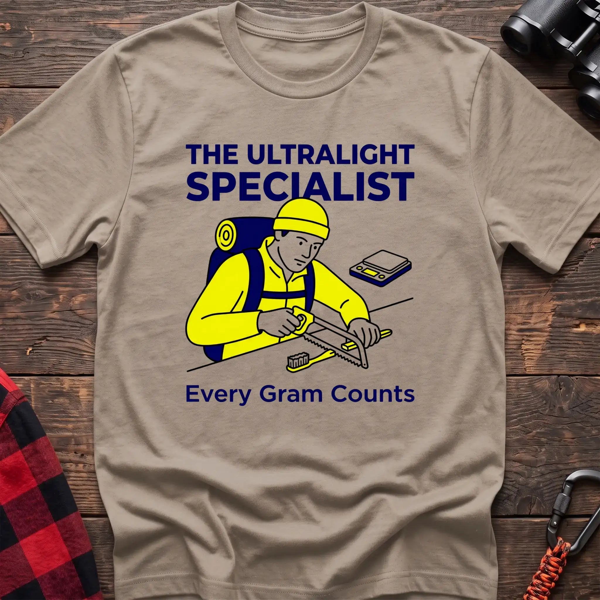 Sand t-shirt with an illustration of a hiker sawing a toothbrush on a table next to a scale, with the text “THE ULTRALIGHT SPECIALIST” and “Every Gram Counts,” laid flat on a wooden surface.