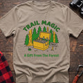 Sand t-shirt with an illustration of an open cooler filled with drinks in a forest setting, with the text “TRAIL MAGIC” and “A Gift From The Forest,” laid flat on a wooden surface.