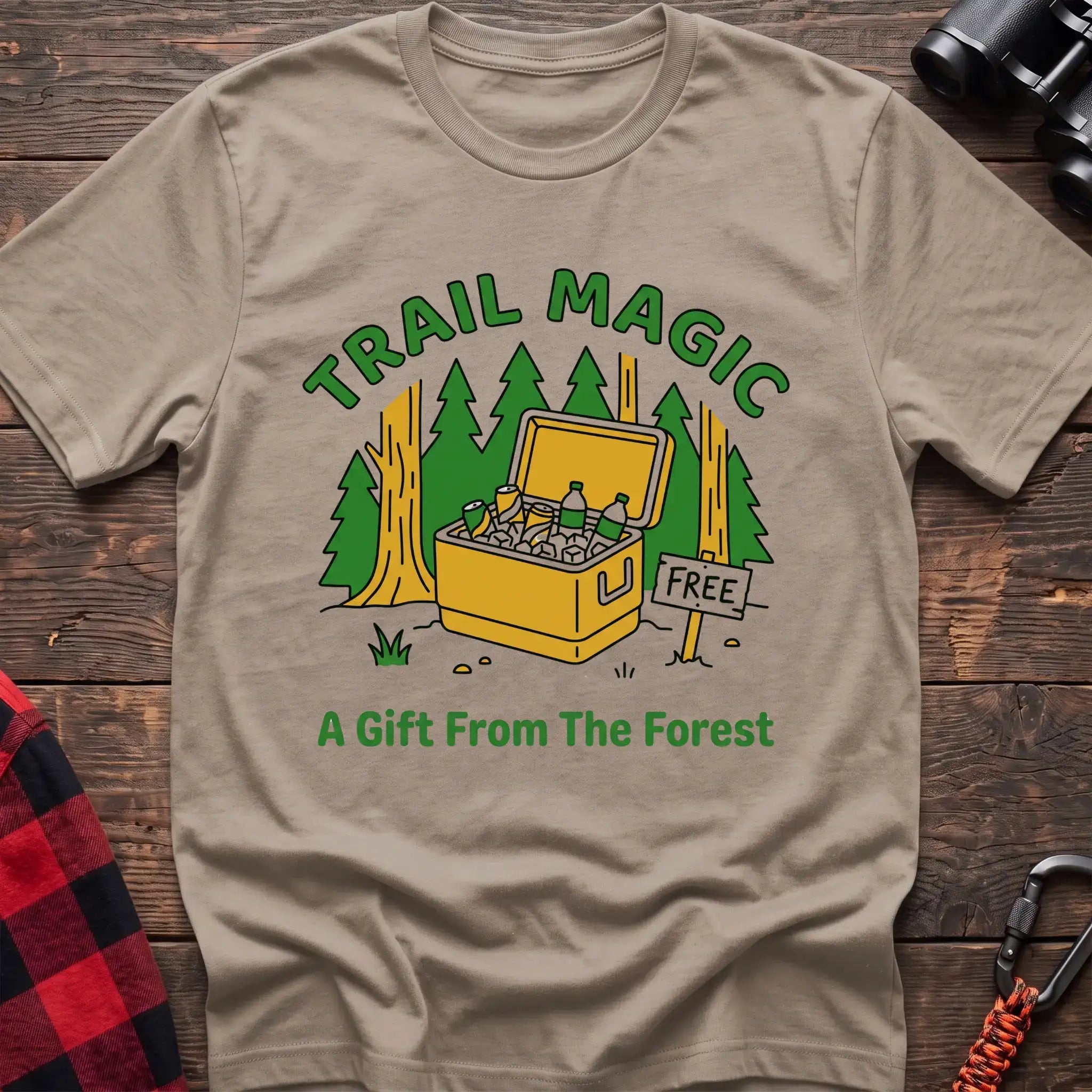Sand t-shirt with an illustration of an open cooler filled with drinks in a forest setting, with the text “TRAIL MAGIC” and “A Gift From The Forest,” laid flat on a wooden surface.
