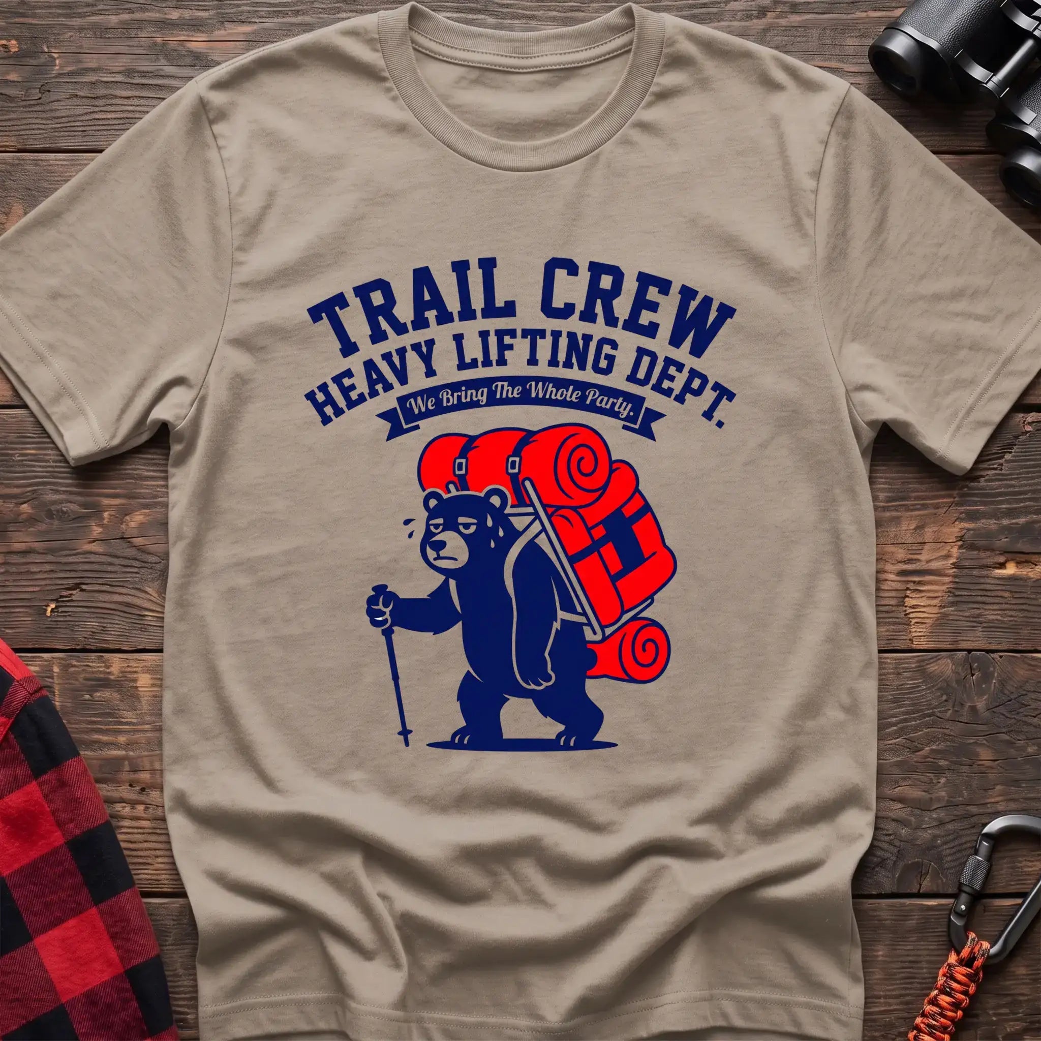Sand t-shirt with a cartoon dark blue bear carrying a large red backpack and hiking pole, with the text “TRAIL CREW HEAVY LIFTING DEPT. We Bring The Whole Party,” laid flat on a wooden surface.