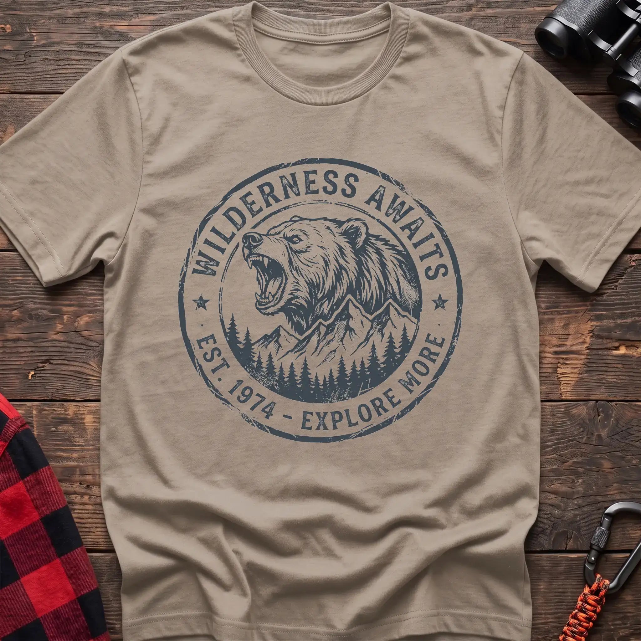 Sand t-shirt with a circular dark gray badge design featuring a roaring bear head above mountains and pine trees, with the text “WILDERNESS AWAITS” and “EST. 1974 - EXPLORE MORE,” laid flat on a wooden surface.