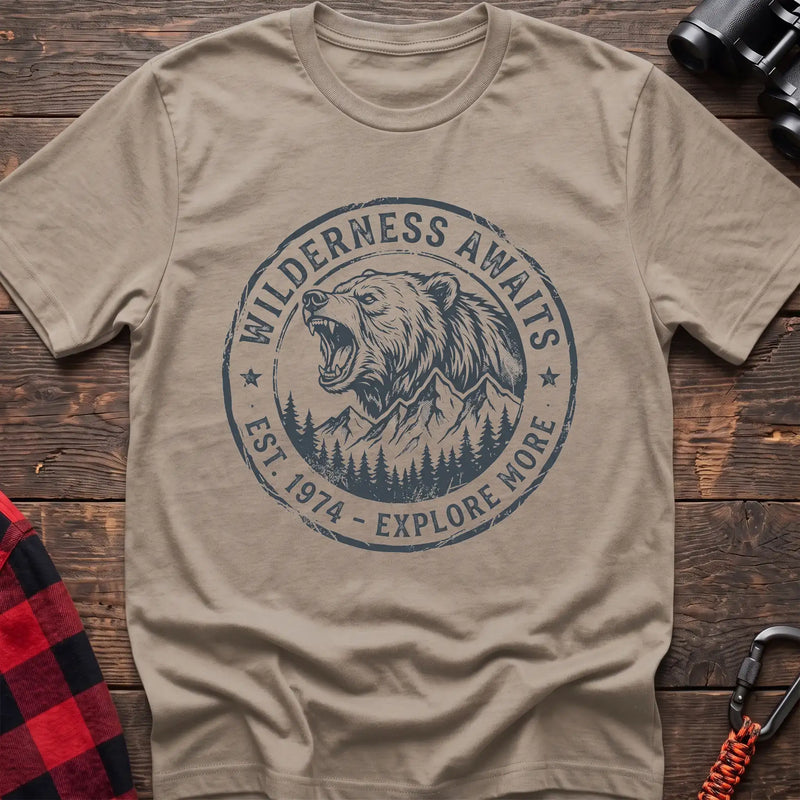 Sand t-shirt with a circular dark gray badge design featuring a roaring bear head above mountains and pine trees, with the text “WILDERNESS AWAITS” and “EST. 1974 - EXPLORE MORE,” laid flat on a wooden surface.