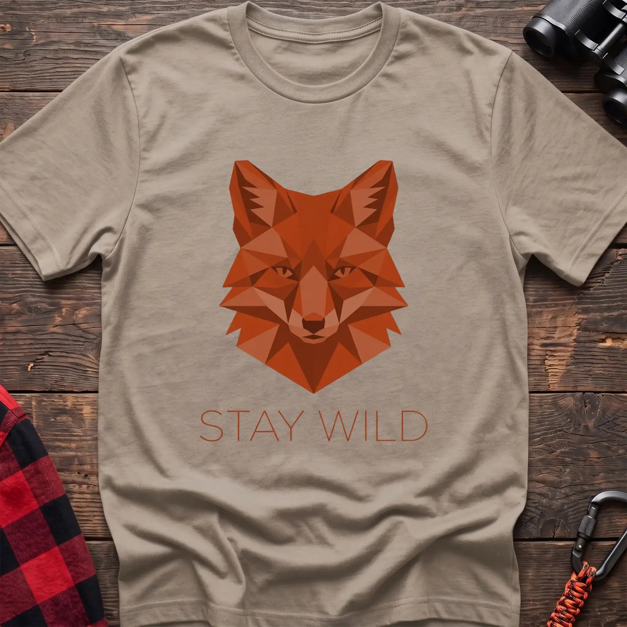 Sand t-shirt with a geometric low-poly orange fox head and the text “STAY WILD” printed below in matching orange, laid flat on a wooden surface.