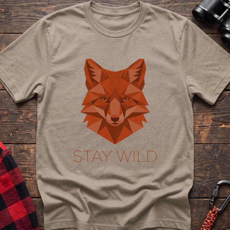 Sand t-shirt with a geometric low-poly orange fox head and the text “STAY WILD” printed below in matching orange, laid flat on a wooden surface.