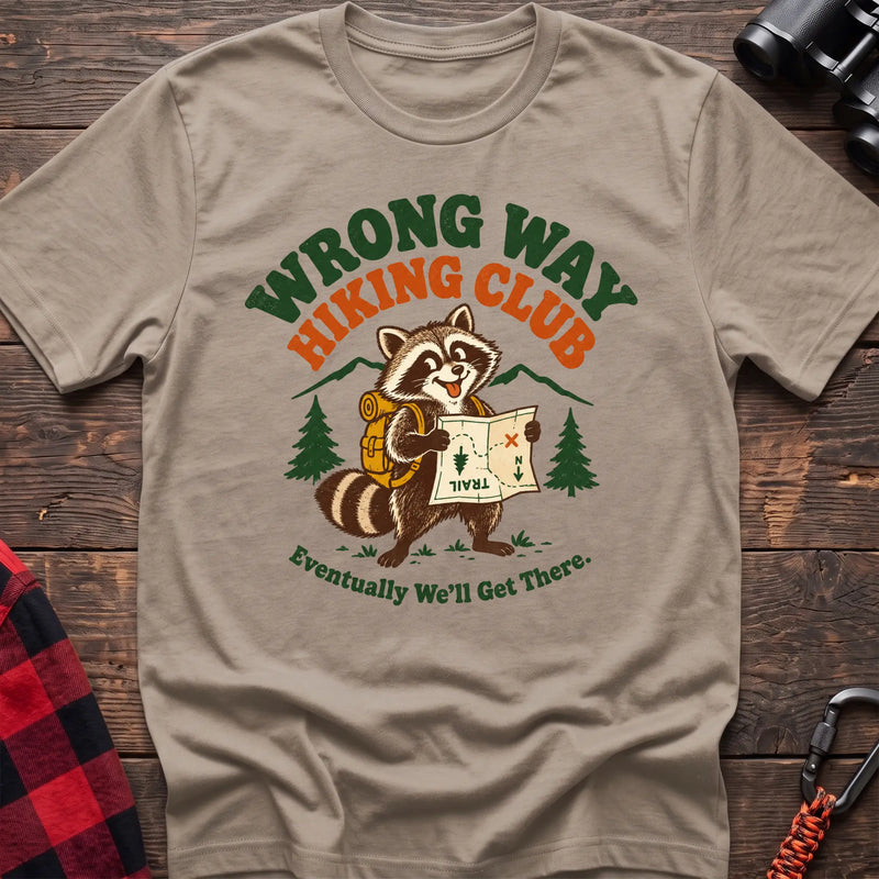 Sand cotton t-shirt with a raccoon holding a map graphic and Wrong Way Hiking Club text on rustic wood