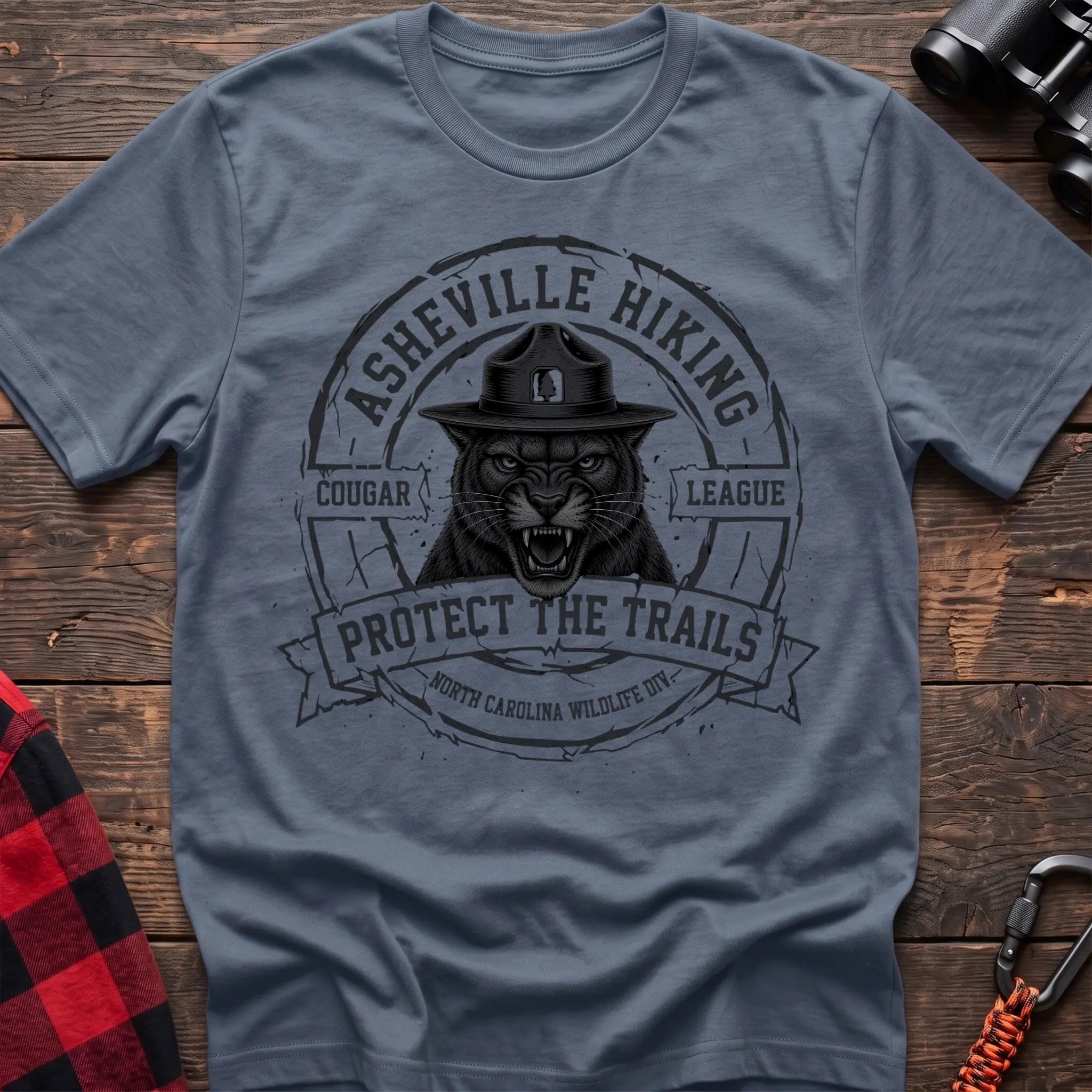Stone blue cotton t-shirt with vintage Asheville Hiking Cougar League graphic of a roaring cougar in a park ranger hat, Protect the Trails North Carolina Wildlife Division badge, flat lay on rustic wood with hiking gear