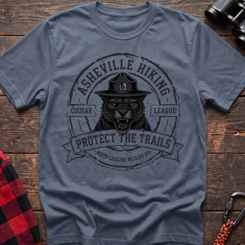Stone blue cotton t-shirt with vintage Asheville Hiking Cougar League graphic of a roaring cougar in a park ranger hat, Protect the Trails North Carolina Wildlife Division badge, flat lay on rustic wood with hiking gear