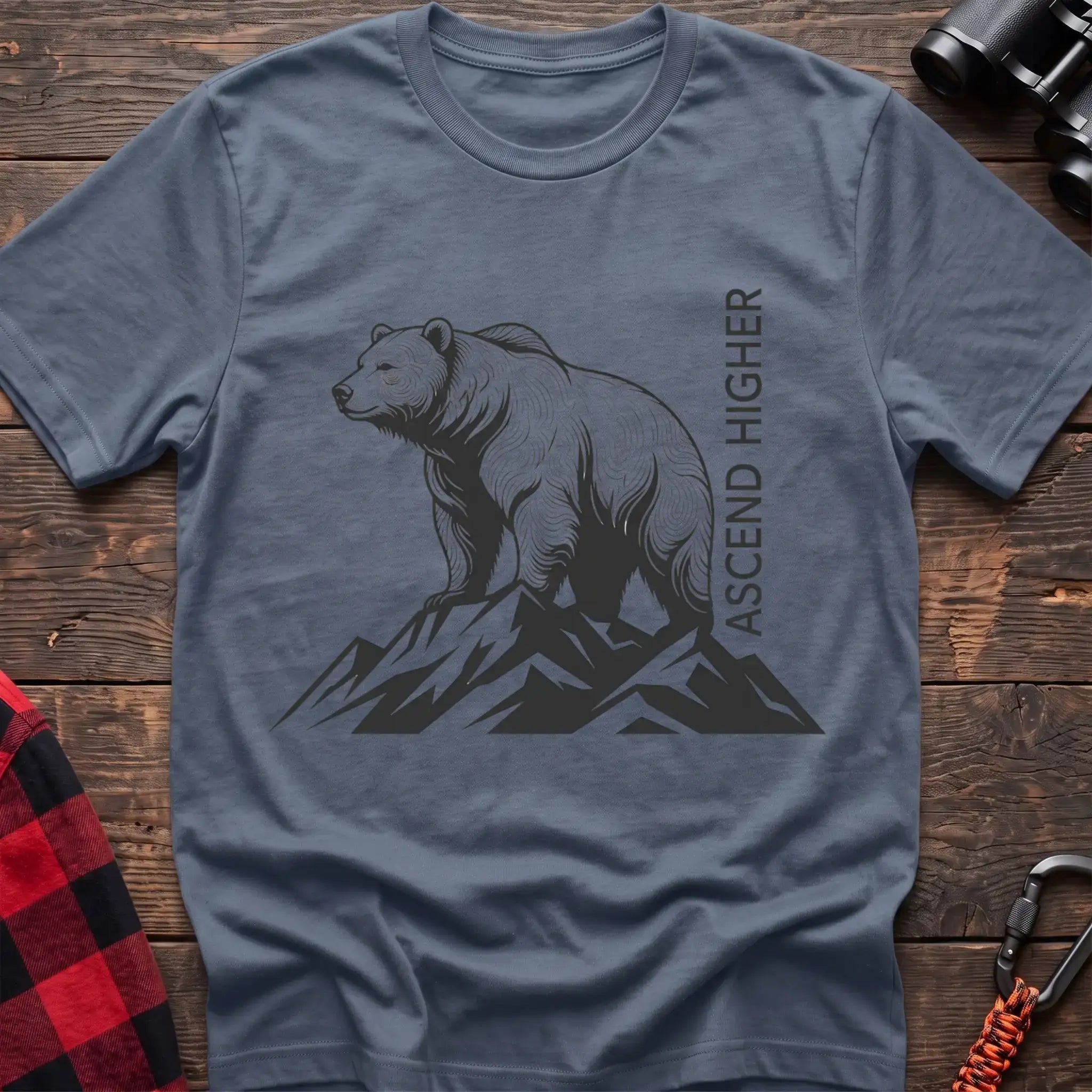 Stone blue t-shirt with a black illustration of a bear standing on mountain peaks, with the vertical text “ASCEND HIGHER” beside it, laid flat on a wooden surface.
