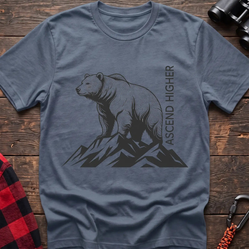 Stone blue t-shirt with a black illustration of a bear standing on mountain peaks, with the vertical text “ASCEND HIGHER” beside it, laid flat on a wooden surface.
