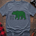 Stone blue t-shirt with a neon green circuit-board patterned silhouette of a walking bear, and the text “DIGITAL DETOX” printed to the left of the design, laid flat on a wooden surface.