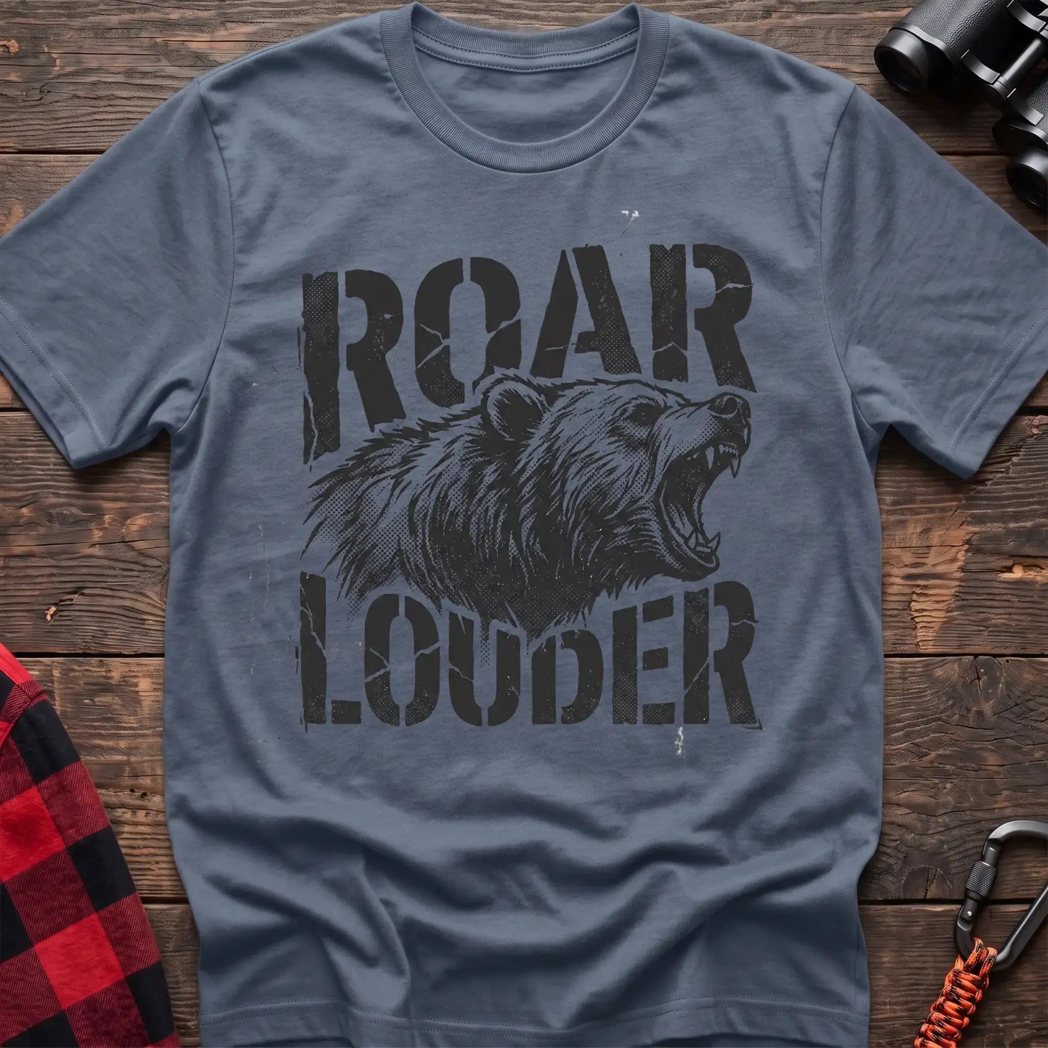 Stone blue t-shirt with a distressed black graphic of a roaring bear head between the bold text “ROAR” and “LOUDER,” laid flat on a wooden surface.