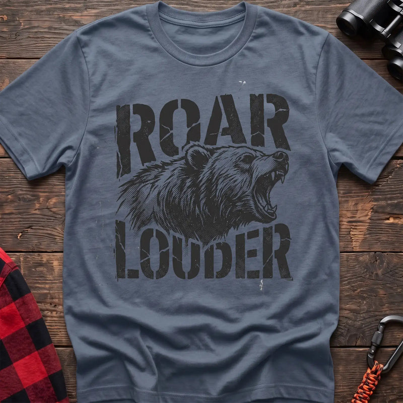 Stone blue t-shirt with a distressed black graphic of a roaring bear head between the bold text “ROAR” and “LOUDER,” laid flat on a wooden surface.