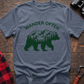 Stone blue t-shirt with a dark green silhouette of a walking bear containing a mountain and pine forest scene inside, with the arched text “WANDER OFTEN” above it, laid flat on a wooden surface.