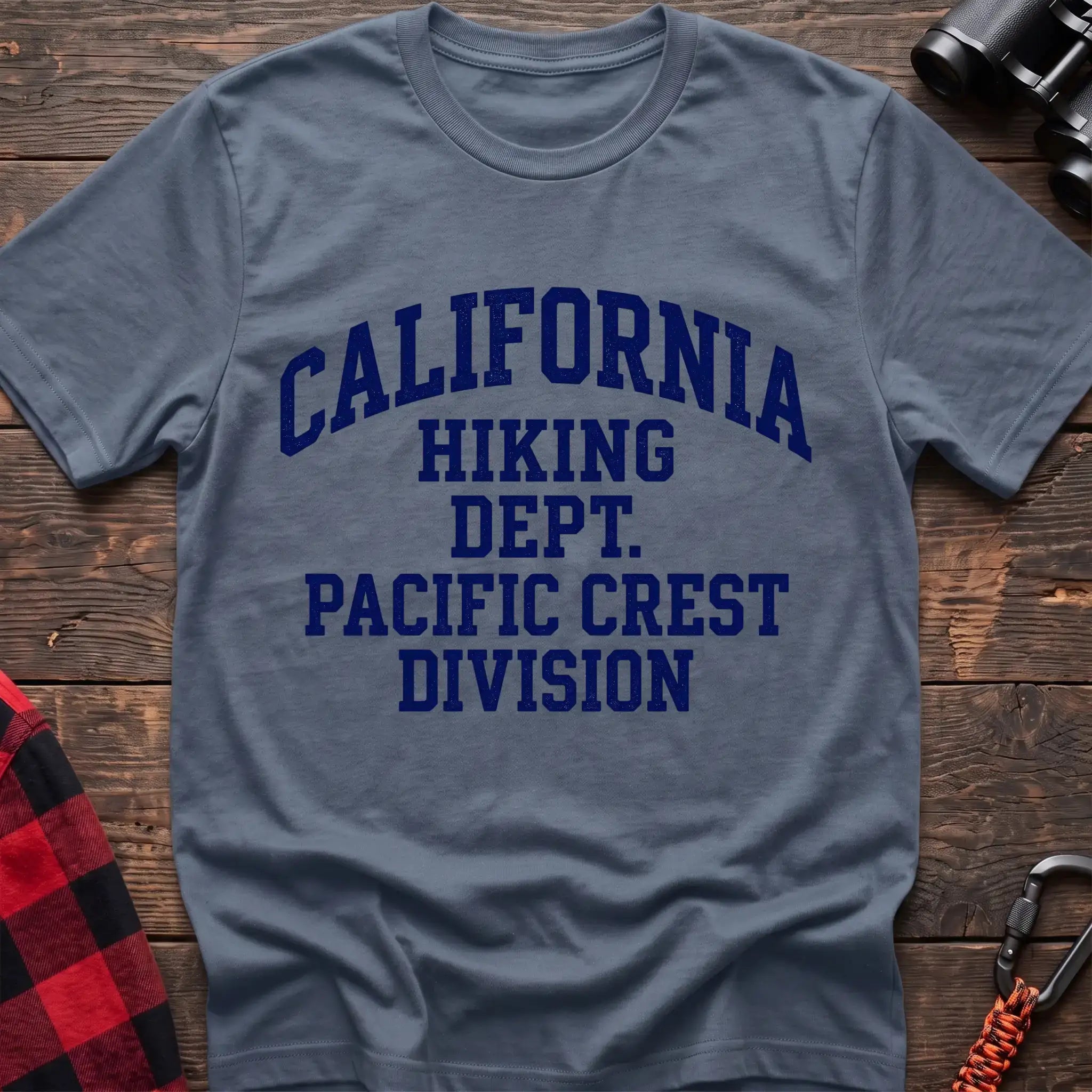 Stone blue t-shirt with distressed blue collegiate-style text reading “CALIFORNIA HIKING DEPT. PACIFIC CREST DIVISION,” laid flat on a wooden surface.