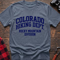 Stone blue t-shirt with distressed dark blue collegiate-style text reading “COLORADO HIKING DEPT. ROCKY MOUNTAIN DIVISION,” laid flat on a wooden surface.
