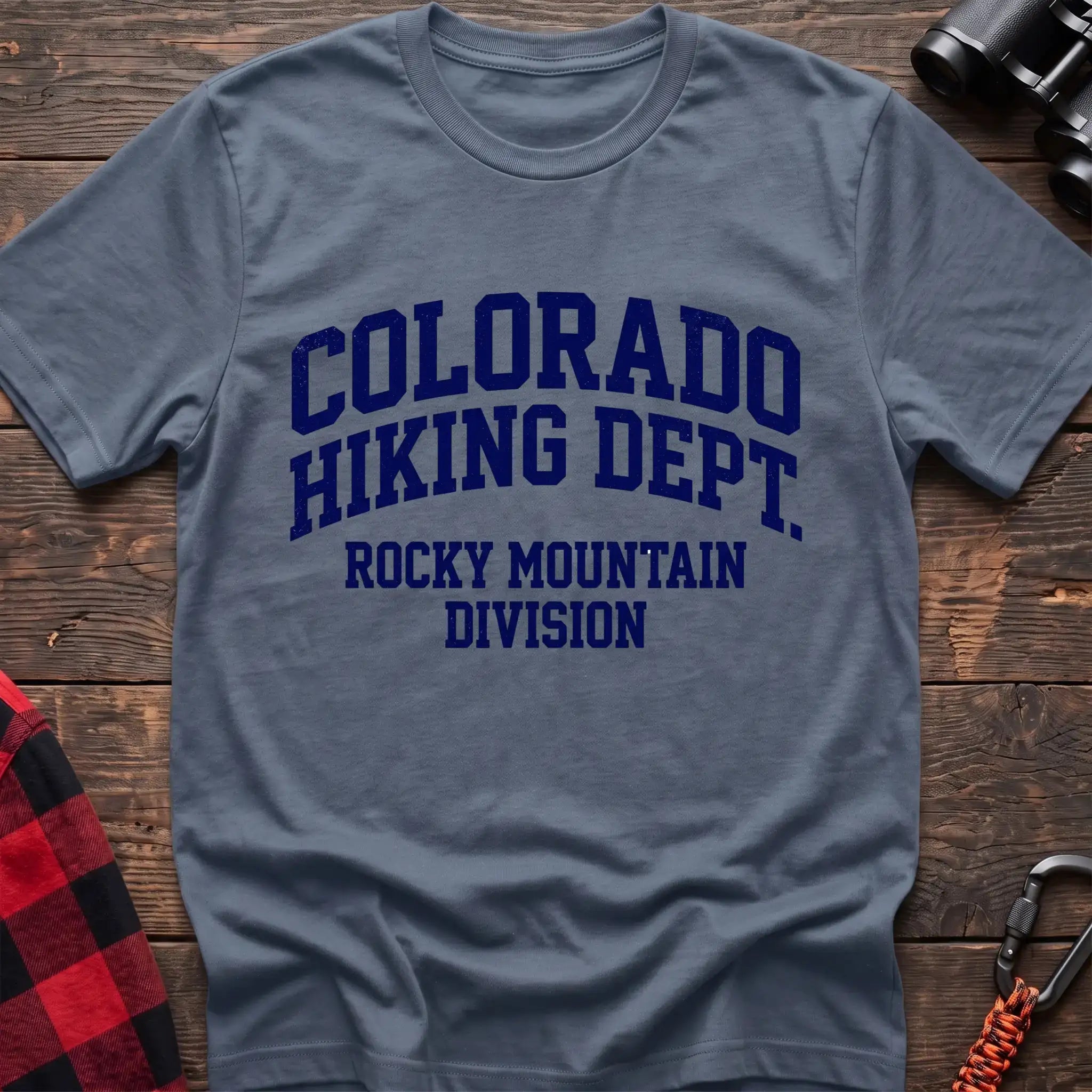 Stone blue t-shirt with distressed dark blue collegiate-style text reading “COLORADO HIKING DEPT. ROCKY MOUNTAIN DIVISION,” laid flat on a wooden surface.