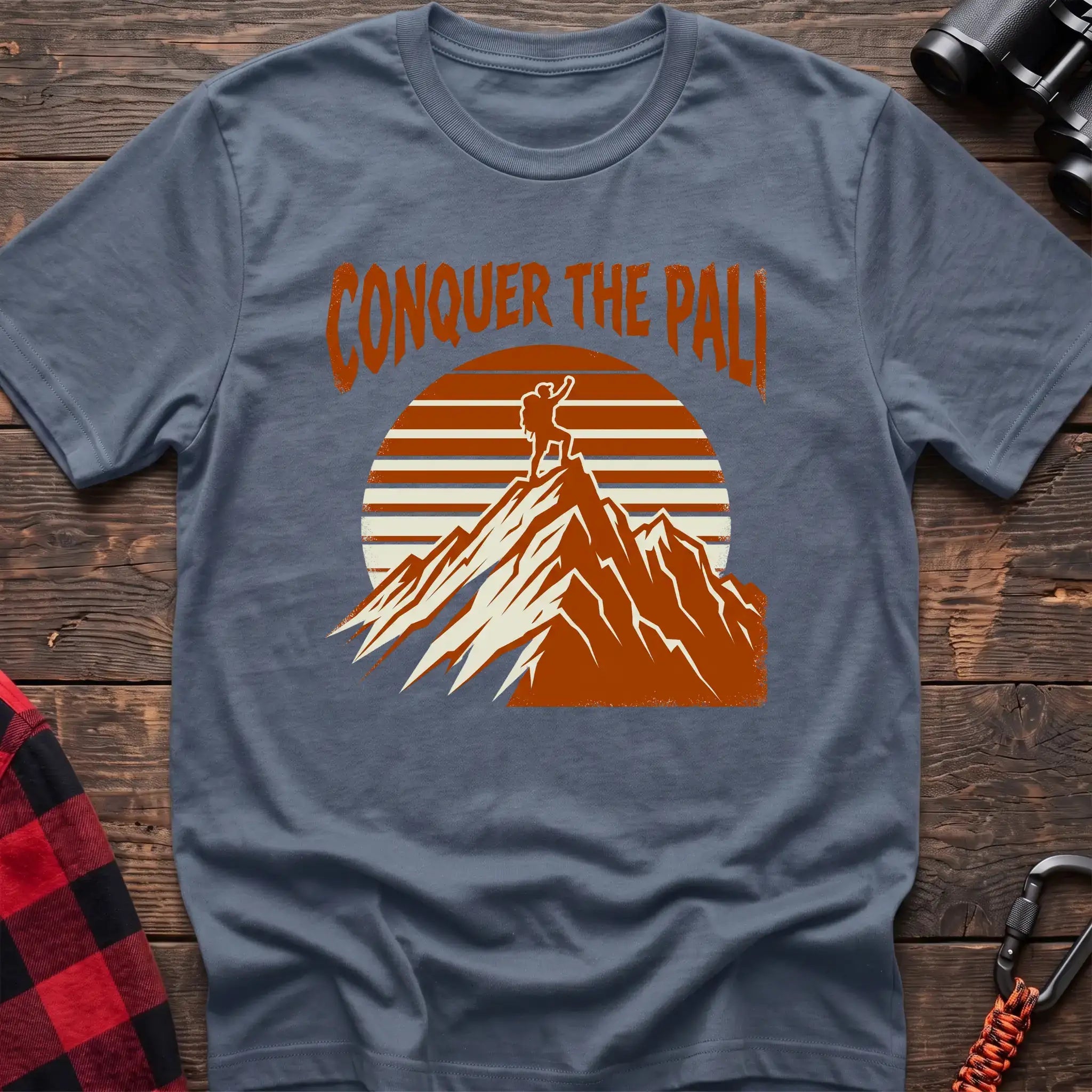 Stone blue t-shirt with orange text reading Conquer the Pali above a hiker standing on a mountain peak with a retro sun graphic