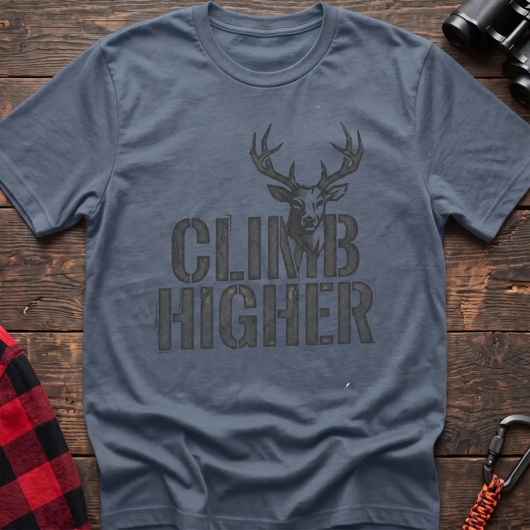 Stone blue t-shirt with a distressed dark graphic of a deer’s head and antlers rising from the bold text “CLIMB HIGHER,” laid flat on a wooden surface.