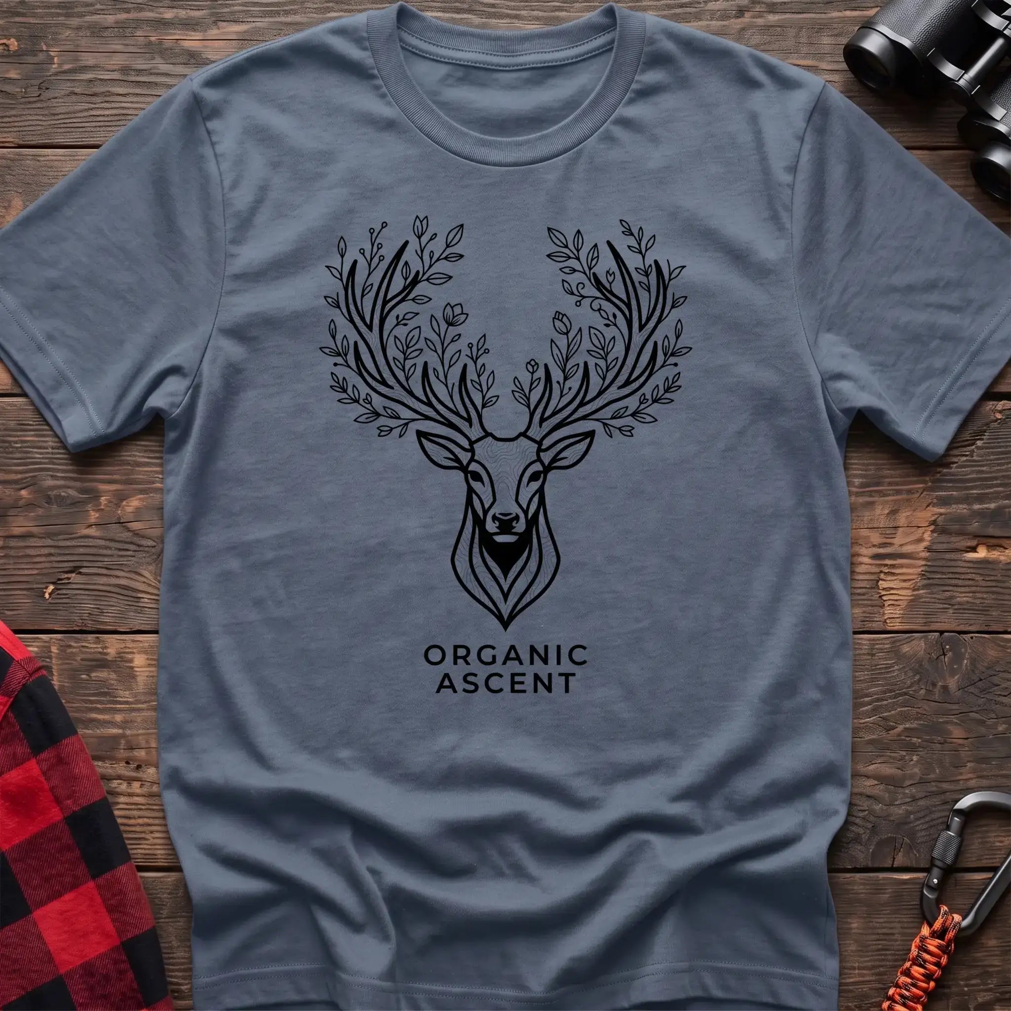 Stone blue t-shirt with a black line drawing of a deer’s head whose antlers are formed from leafy branches and flowers, with the text “ORGANIC ASCENT” printed below, laid flat on a wooden surface.