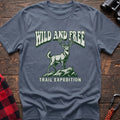 Stone blue t-shirt with a rustic graphic of a deer standing on a rocky outcrop, with the arched text “WILD AND FREE” above and “TRAIL EXPEDITION” below, laid flat on a wooden surface.
