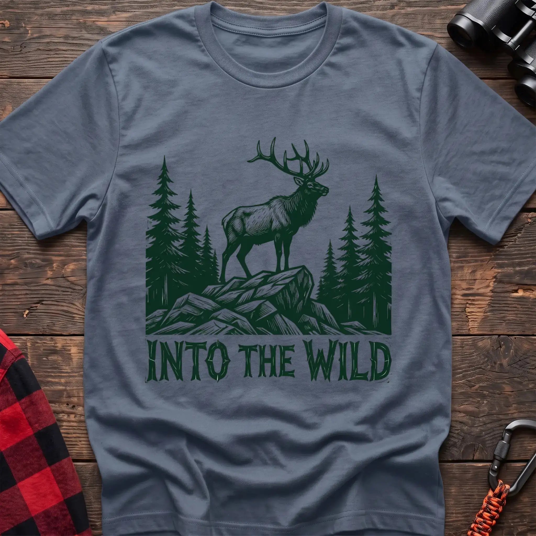 Stone blue t-shirt with a dark green illustration of an elk standing on a rocky outcrop among pine trees, with the text “INTO THE WILD” printed below, laid flat on a wooden surface.
