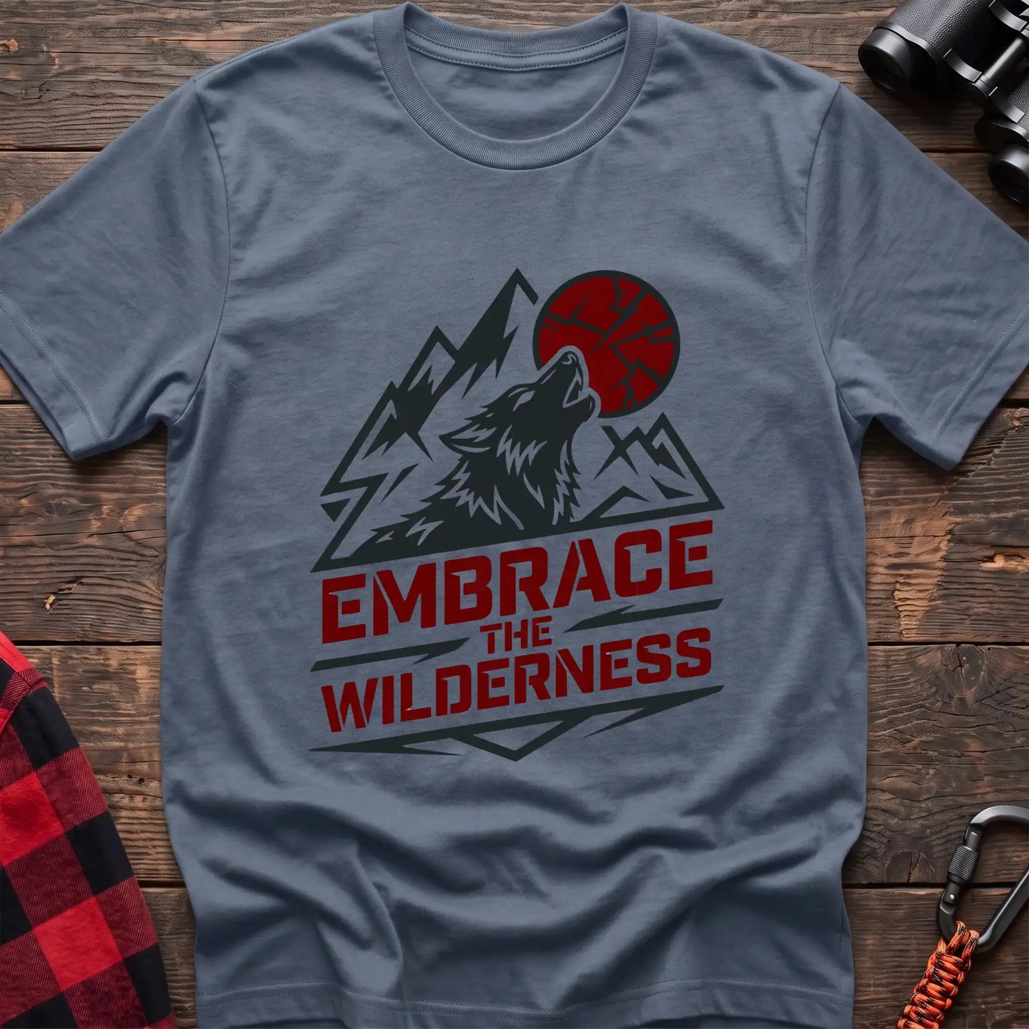 Stone blue t-shirt with a dark graphic of a howling wolf against mountain peaks and a red sun, with the bold red text “EMBRACE THE WILDERNESS” below, laid flat on a wooden surface.