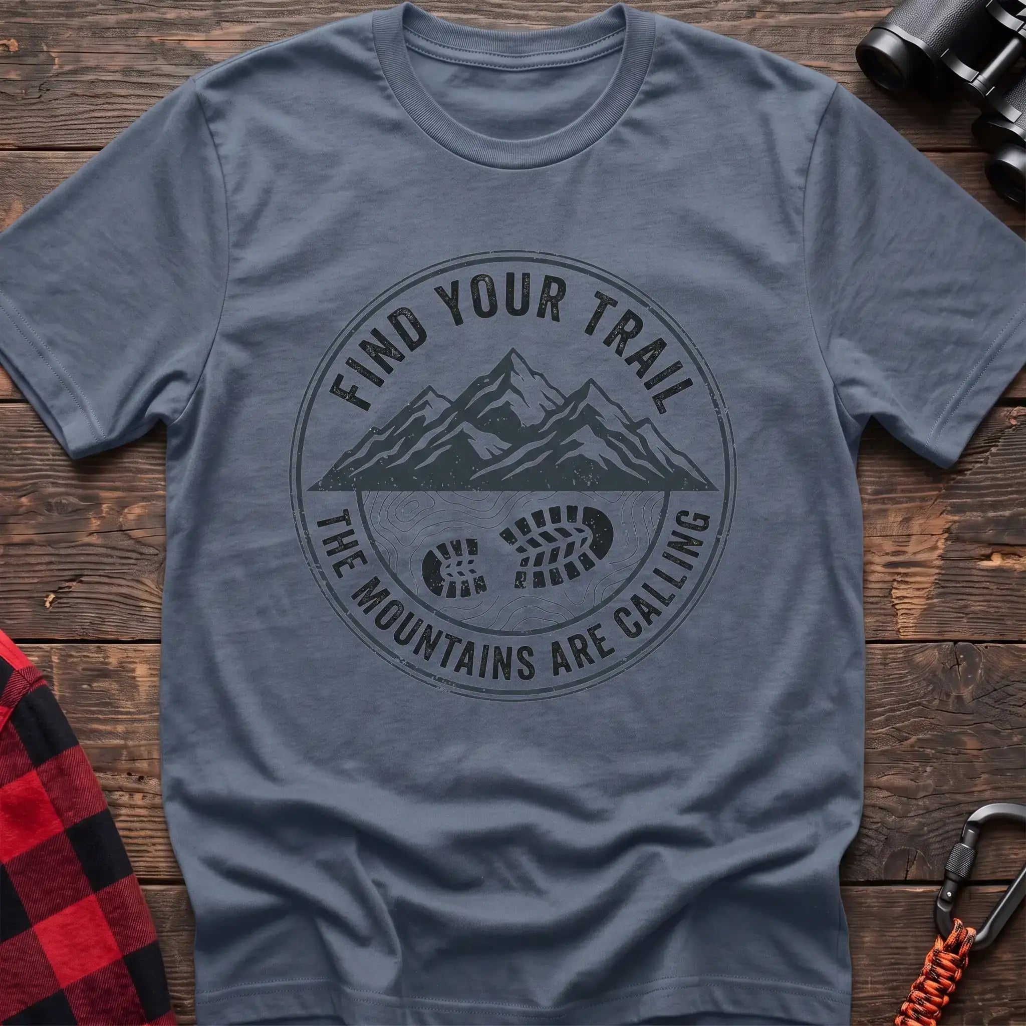 Stone blue t-shirt with a circular dark gray badge design featuring mountain peaks above a hiking boot print on topographic lines, with the text “FIND YOUR TRAIL” and “THE MOUNTAINS ARE CALLING,” laid flat on a wooden surface.