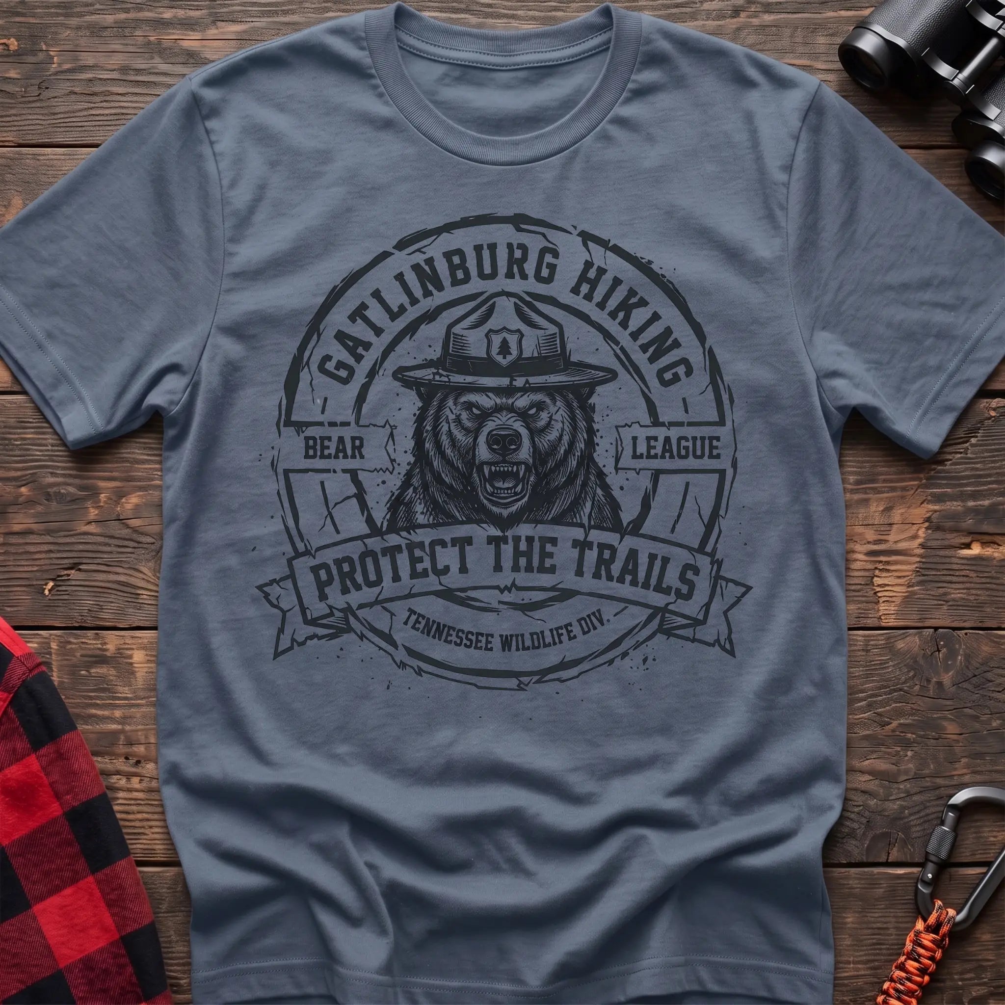 Stone blue cotton t-shirt with vintage Gatlinburg Hiking Bear League graphic of a roaring bear in a park ranger hat, Protect the Trails Tennessee Wildlife Division badge, flat lay on rustic wood with hiking gear