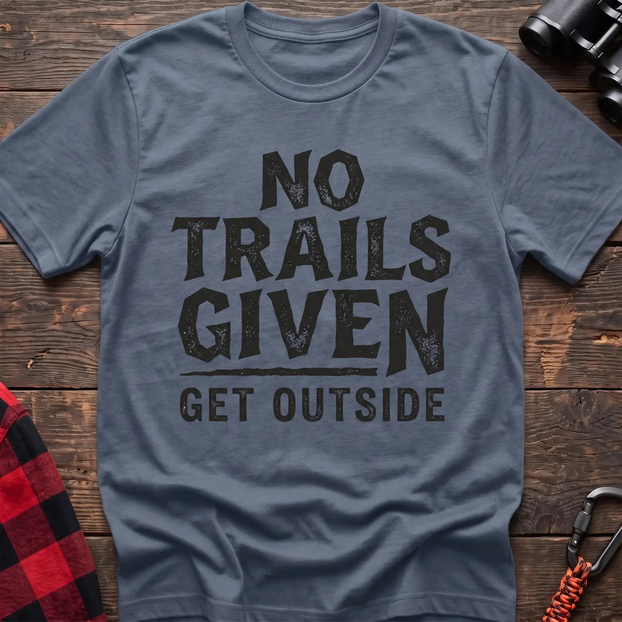 Stone blue t-shirt with distressed black text reading “NO TRAILS GIVEN” and “GET OUTSIDE” below it, laid flat on a wooden surface.