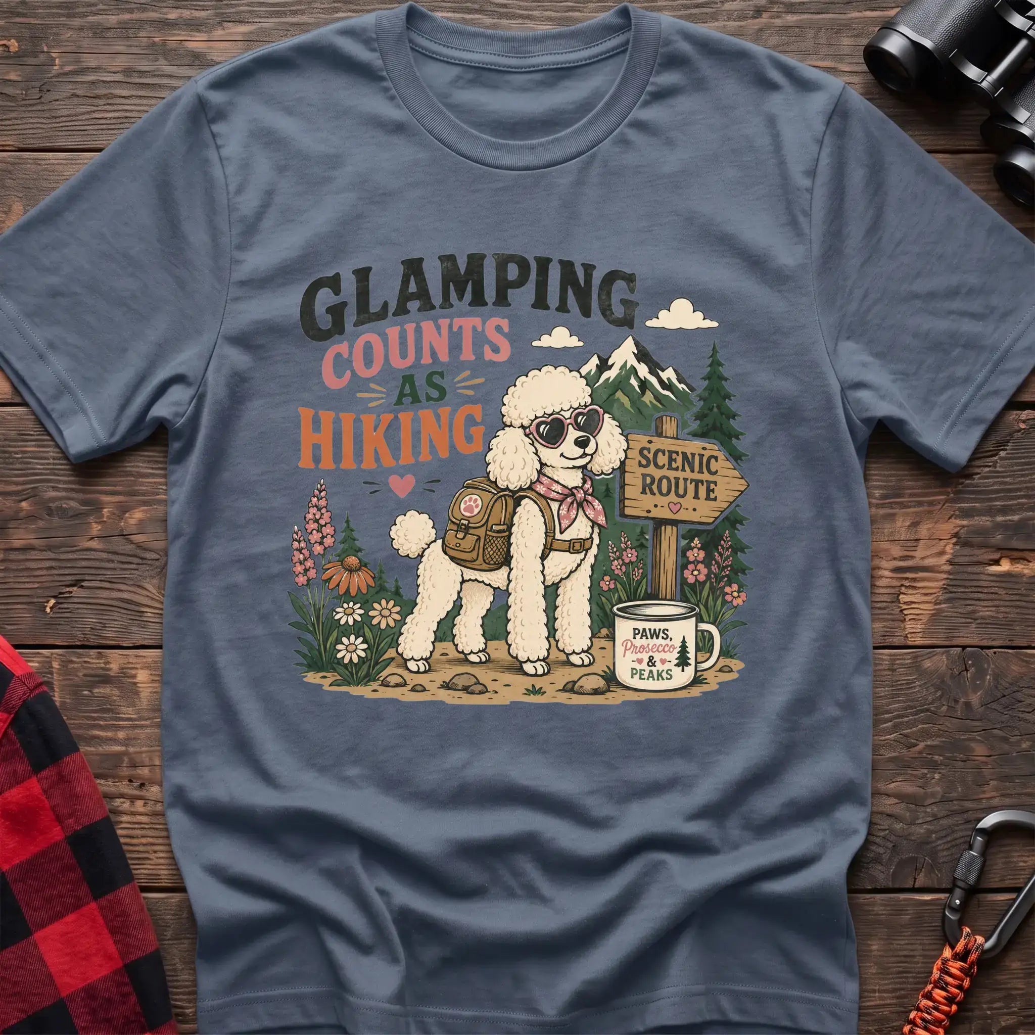 Stone blue t-shirt with a colorful illustration of a poodle in sunglasses and a backpack on a scenic trail, with the text “GLAMPING COUNTS AS HIKING,” laid flat on a wooden surface.