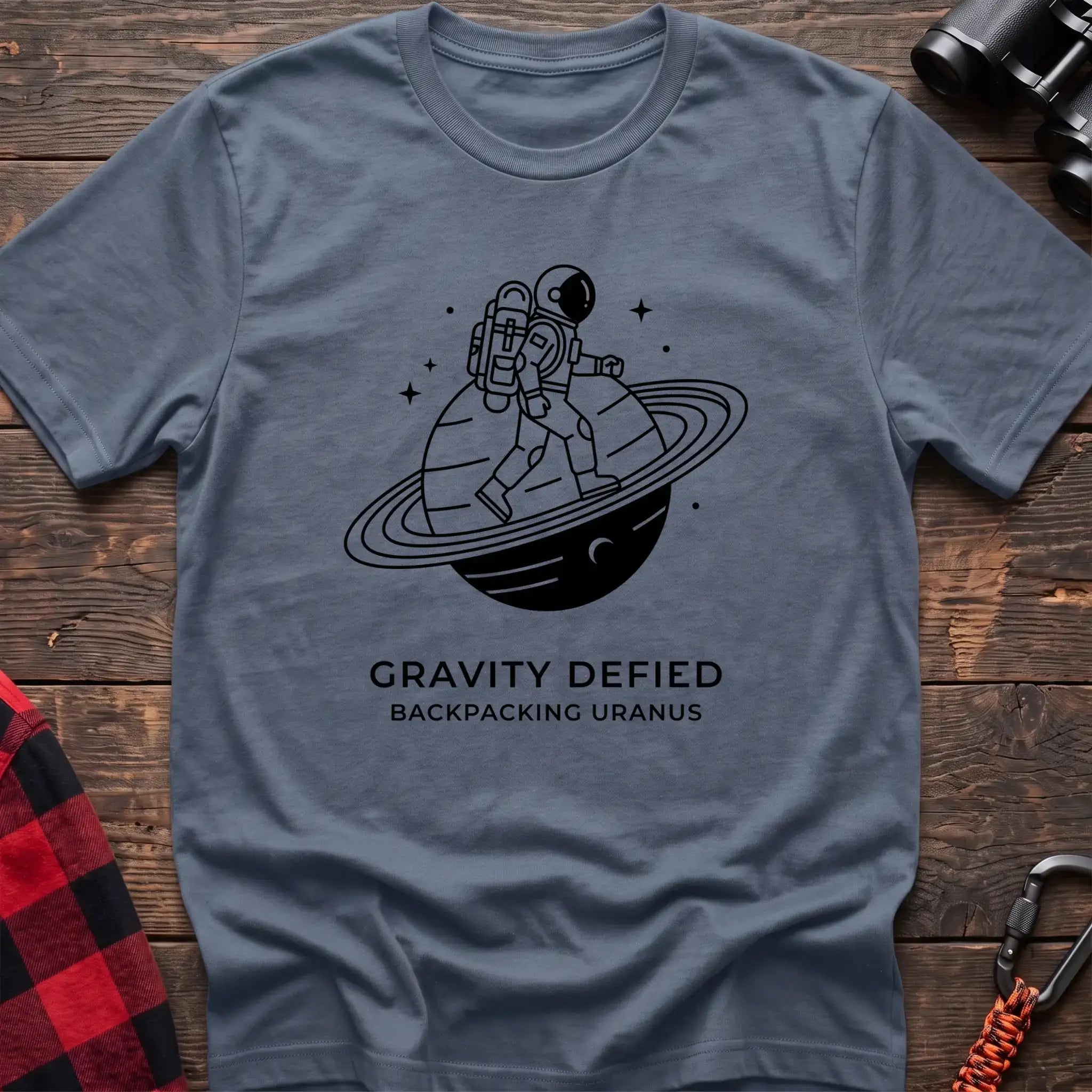 Stone blue crew-neck t-shirt with black line-art graphic of an astronaut backpacking across a ringed planet surrounded by stars, with the text “GRAVITY DEFIED / BACKPACKING URANUS” printed below, laid flat on a wooden surface.