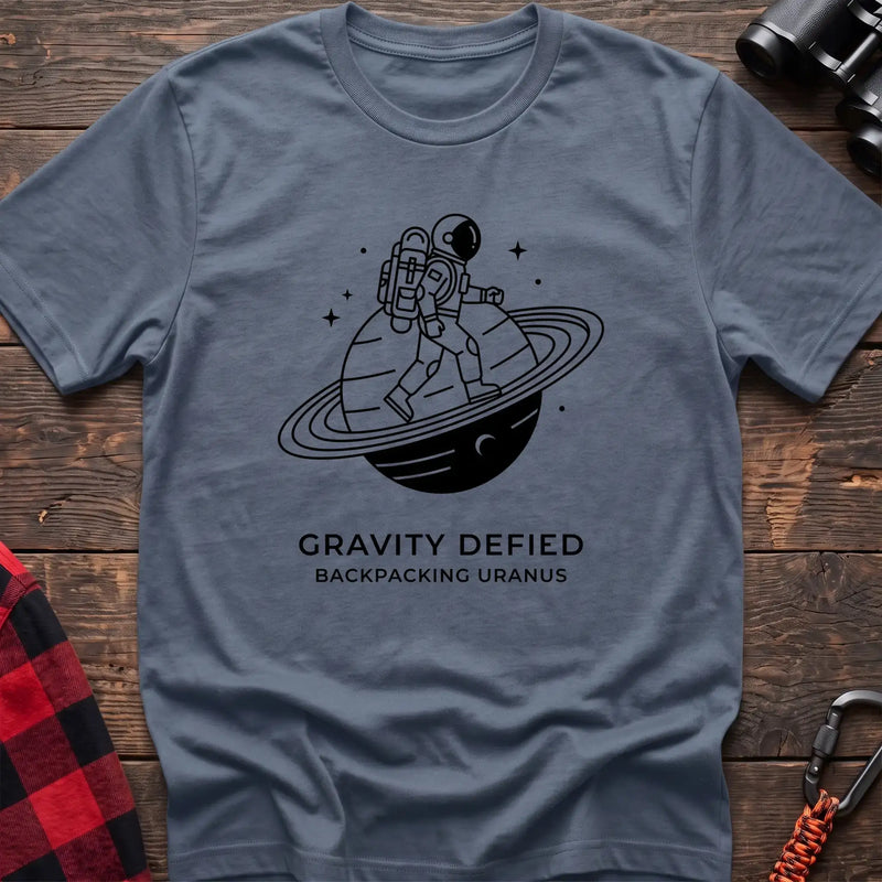 Stone blue crew-neck t-shirt with black line-art graphic of an astronaut backpacking across a ringed planet surrounded by stars, with the text “GRAVITY DEFIED / BACKPACKING URANUS” printed below, laid flat on a wooden surface.