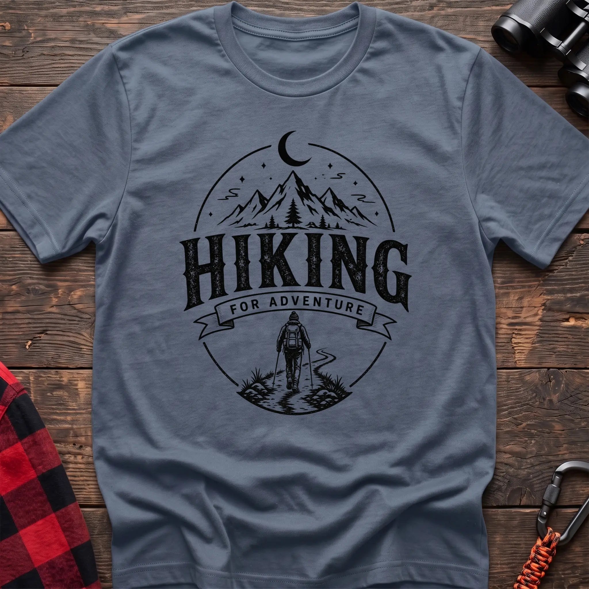 Stone blue cotton t-shirt with vintage Hiking for Adventure graphic of mountains under a crescent moon and a hiker on a trail, flat lay on rustic wood with hiking gear