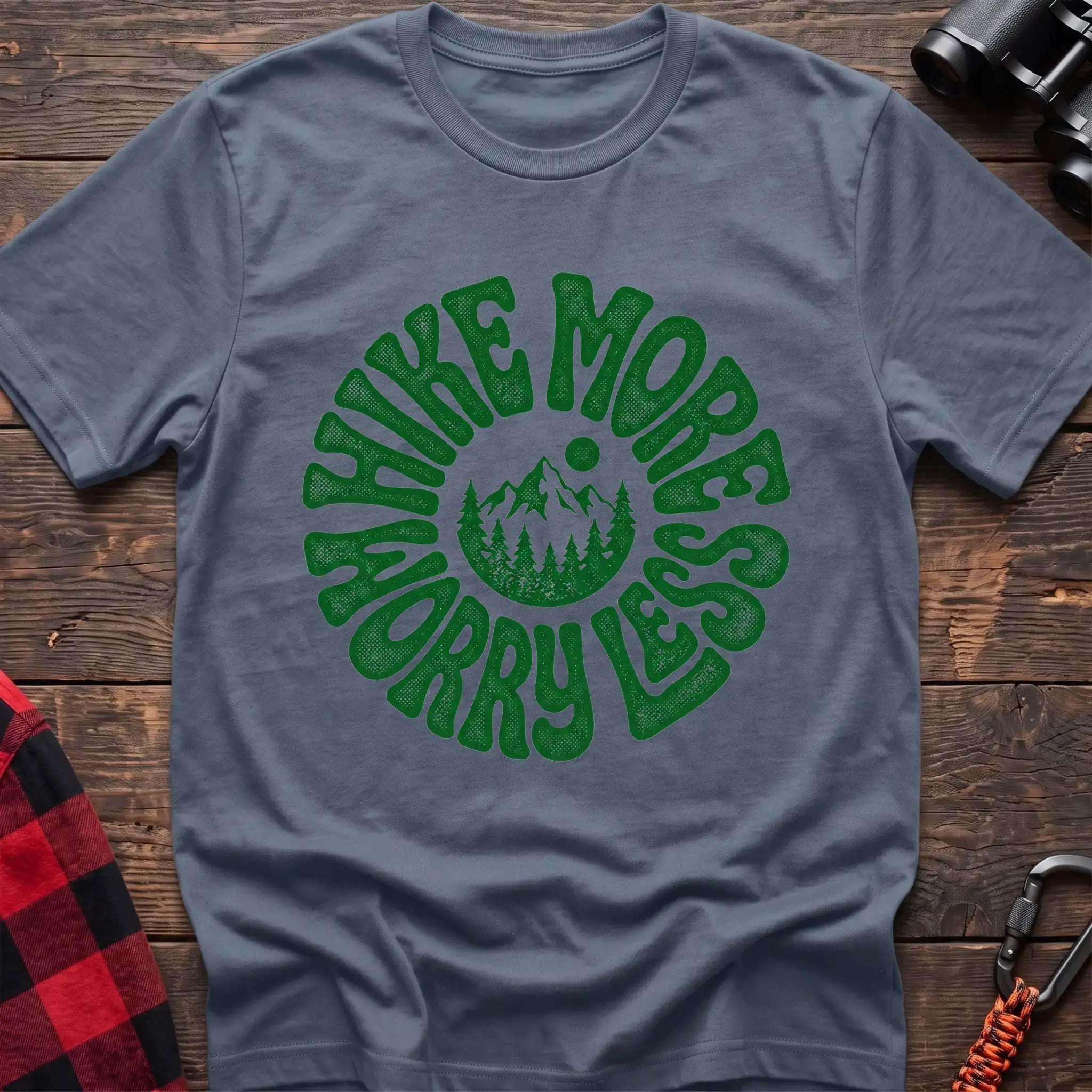 Stone blue t-shirt with a circular green design featuring mountains and pine trees in the center, surrounded by the text “HIKE MORE WORRY LESS,” laid flat on a wooden surface.