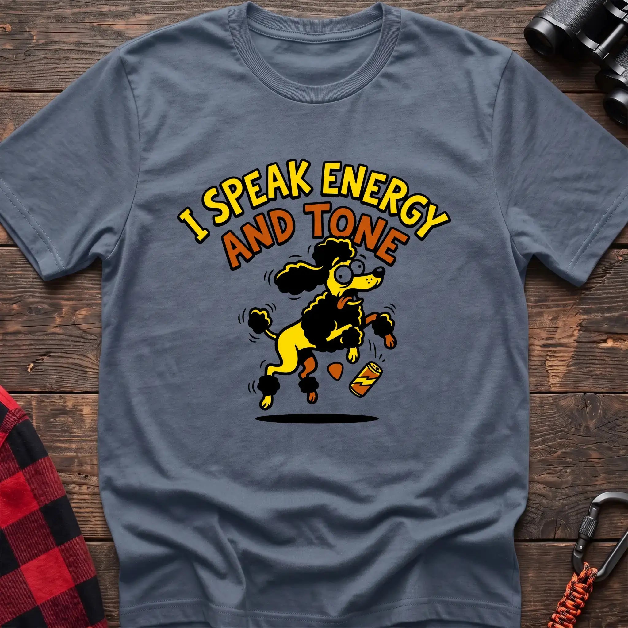 Stone blue t-shirt with a cartoon yellow and black poodle jumping excitedly next to a falling energy drink can, with the arched text “I SPEAK ENERGY AND TONE,” laid flat on a wooden surface.