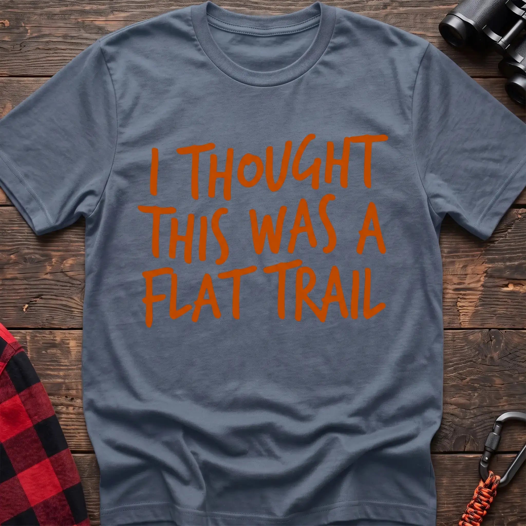 Stone blue t-shirt with bold orange handwritten-style text reading “I THOUGHT THIS WAS A FLAT TRAIL,” laid flat on a wooden surface.