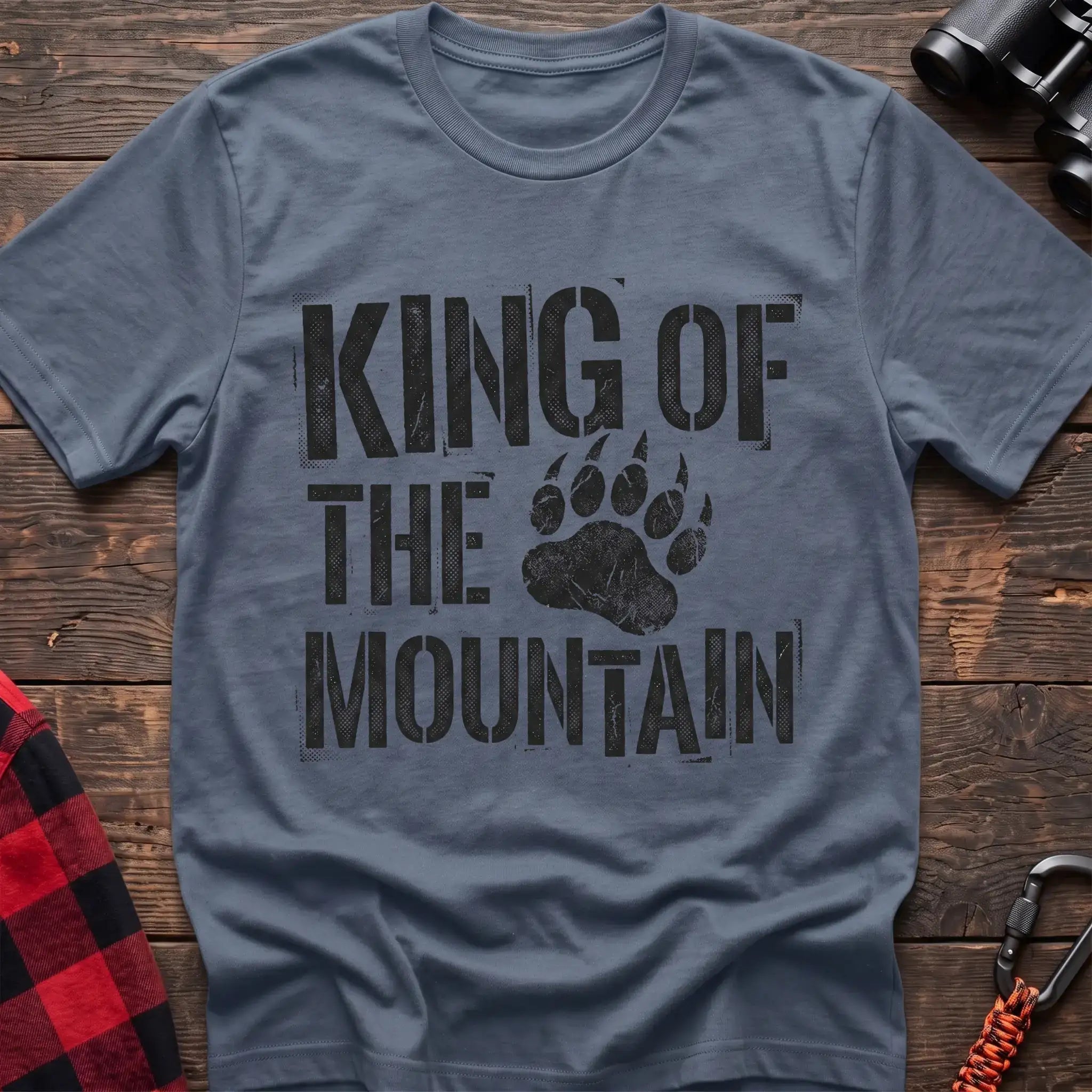 Stone blue t-shirt with distressed black text reading “KING OF THE MOUNTAIN” and a large bear paw print integrated into the design, laid flat on a wooden surface.