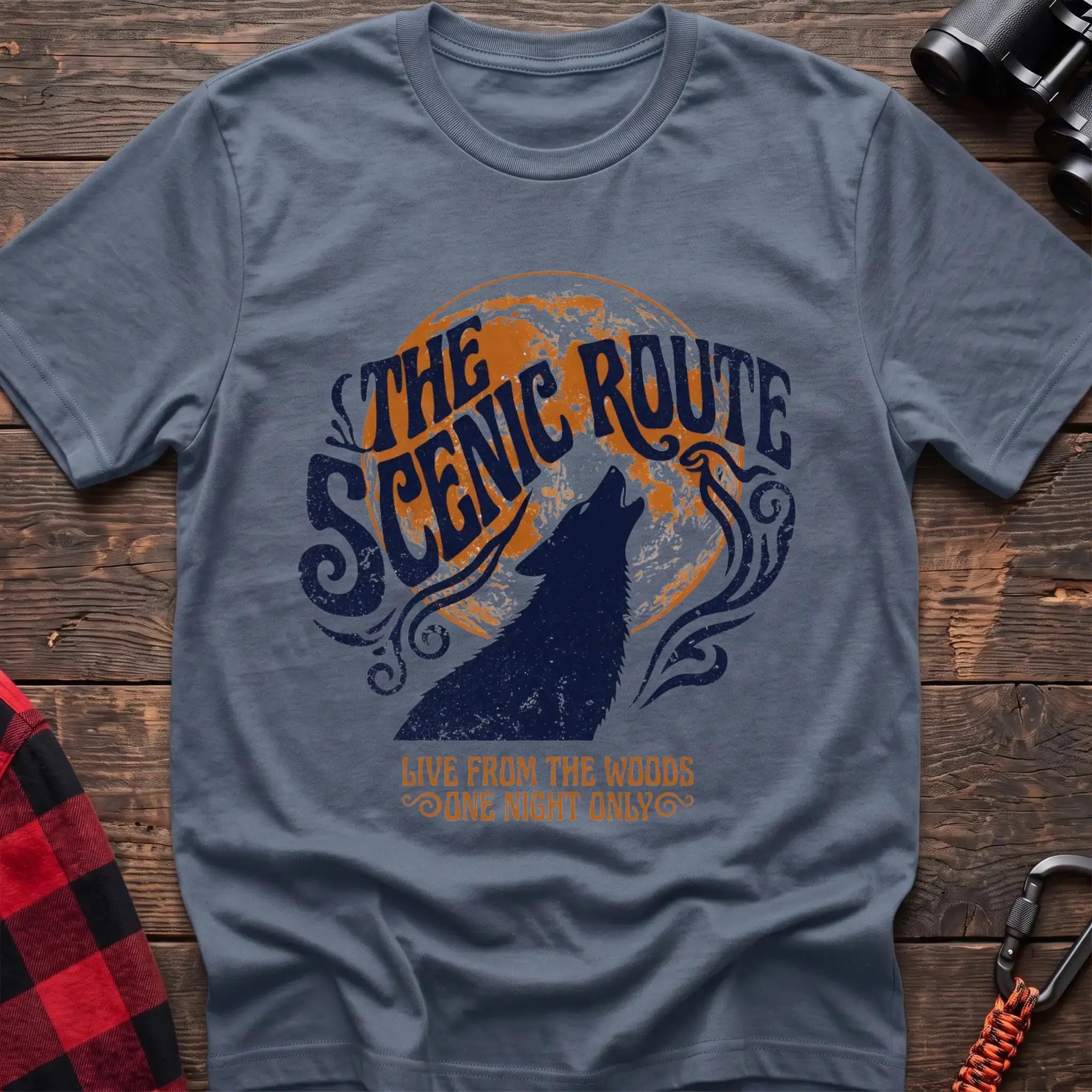 Stone blue t-shirt with a dark blue howling wolf silhouette against an orange circle, with the arched text “THE SCENIC ROUTE” above and “LIVE FROM THE WOODS / ONE NIGHT ONLY” below, laid flat on a wooden surface.