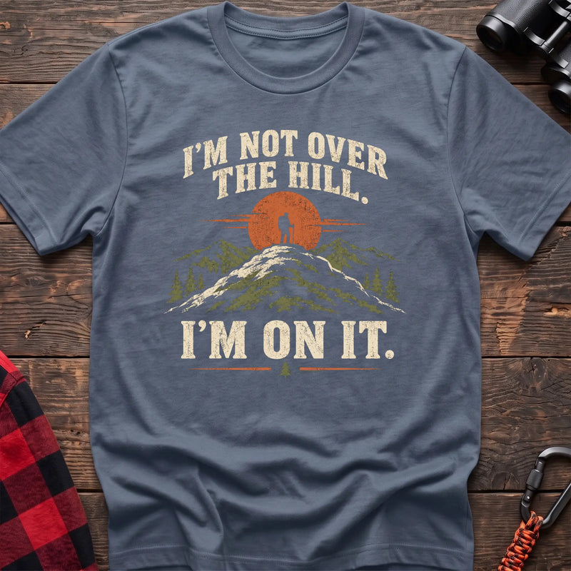 Stone blue cotton t-shirt with a hiker on a summit graphic and I’m Not Over the Hill I’m On It text on rustic wood